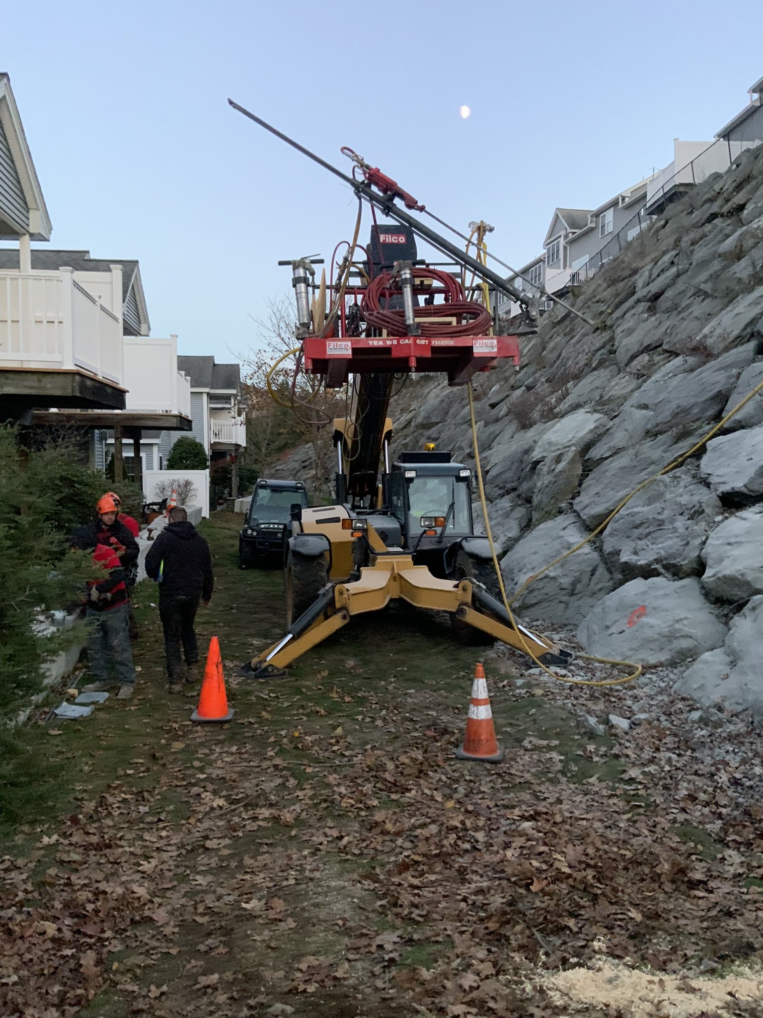 Construction workers and equipment on a pathway next to a rocky hillside, with a crane and orange safety cones, under a clear sky with the moon visible.