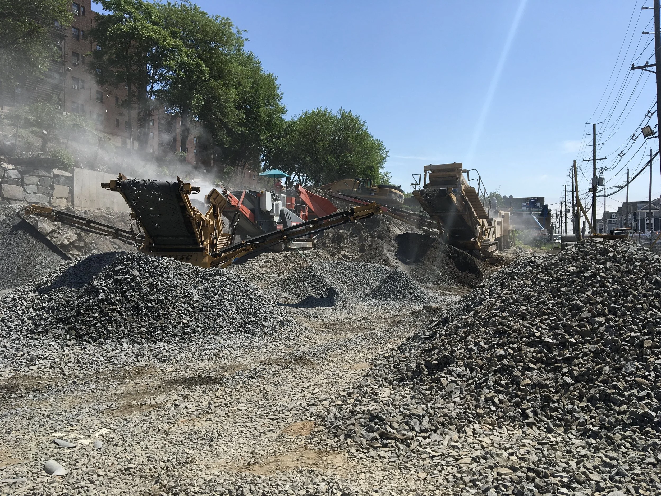 Construction site with heavy machinery working on a dirt and gravel road repair, high from a blue sky and trees in the background.
