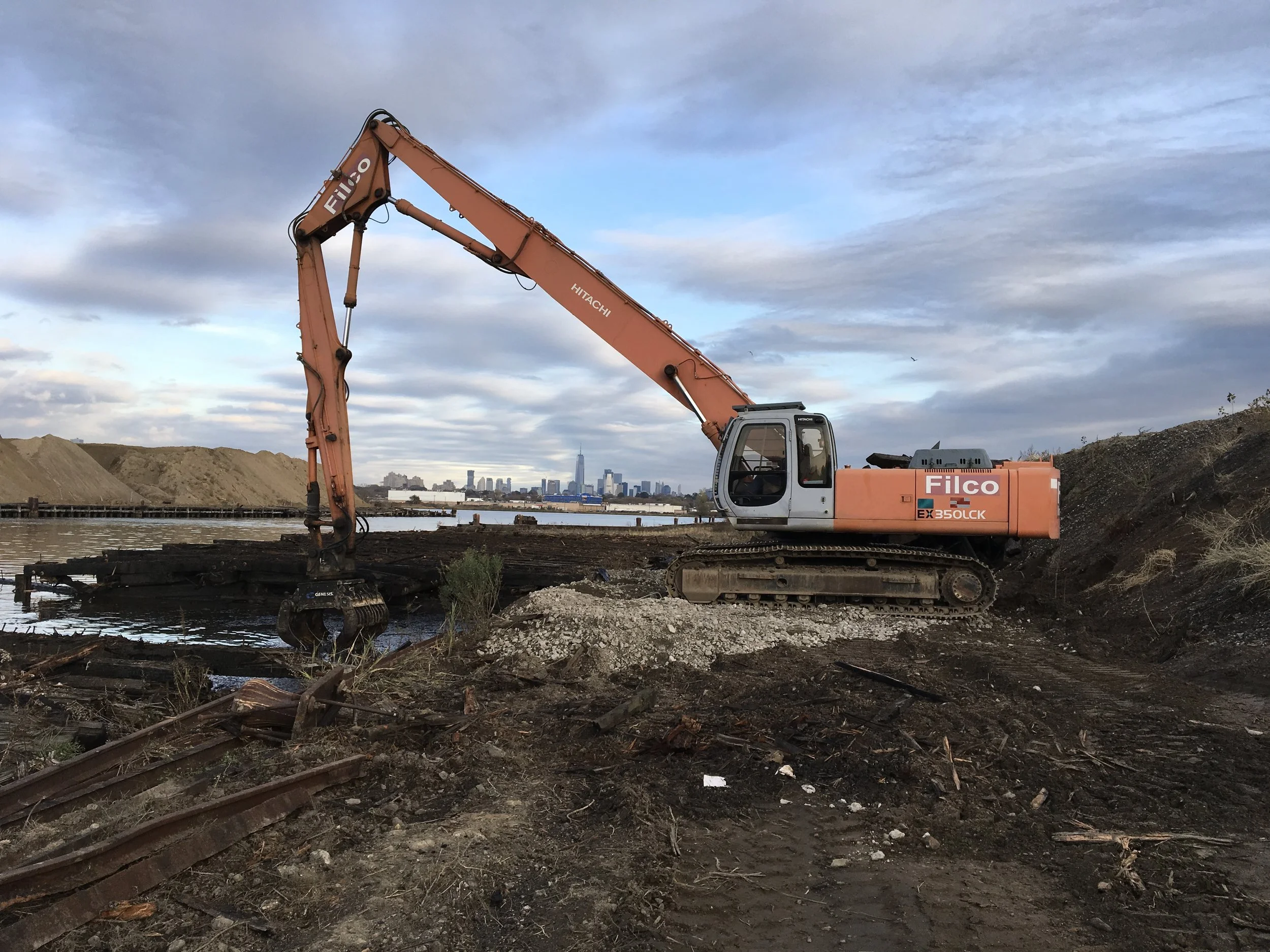 An orange and gray excavator at a construction site near water with a city skyline in the background.