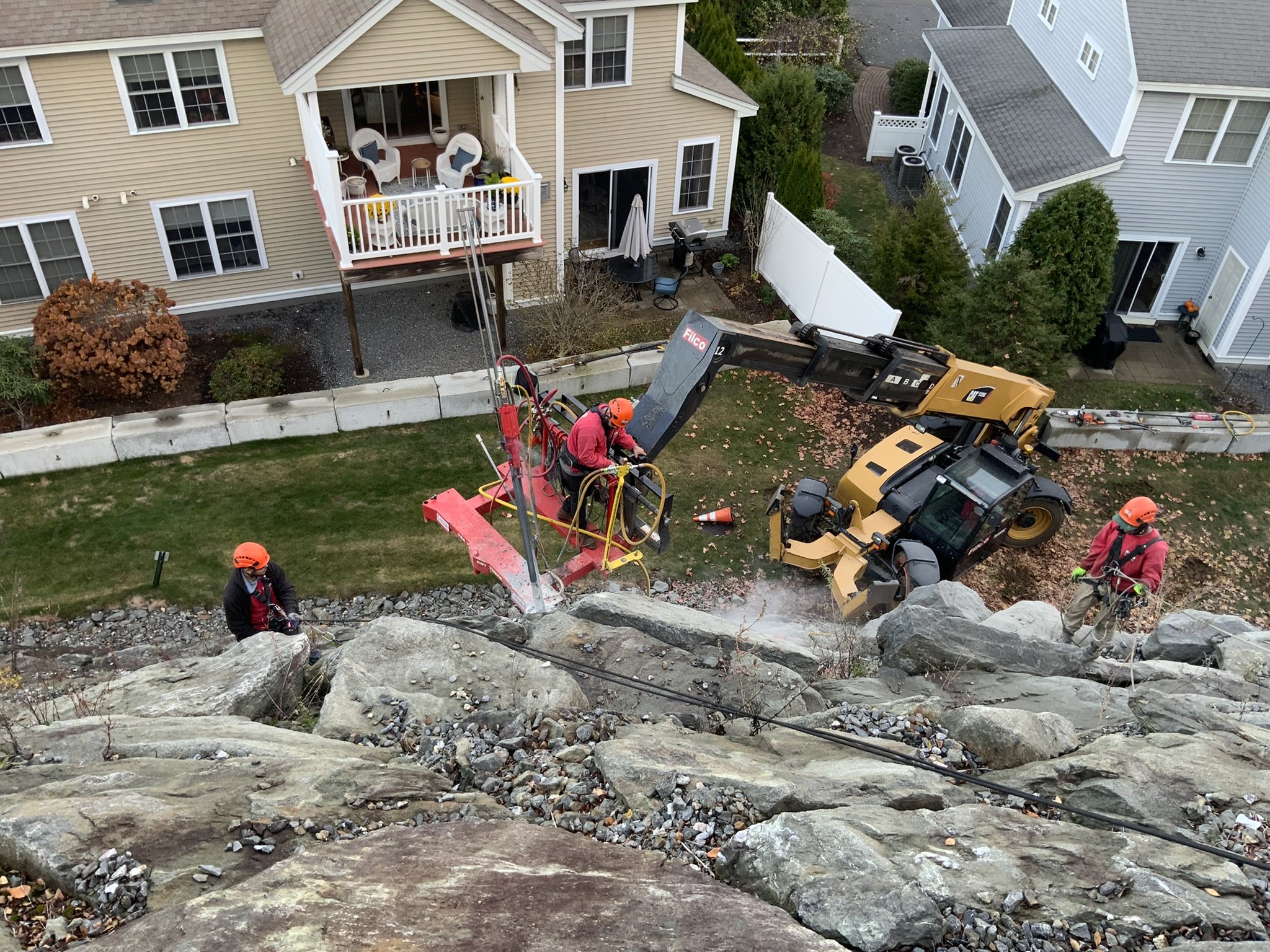 Workers in safety gear using heavy machinery and tools to perform rock or cliff work near residential houses.