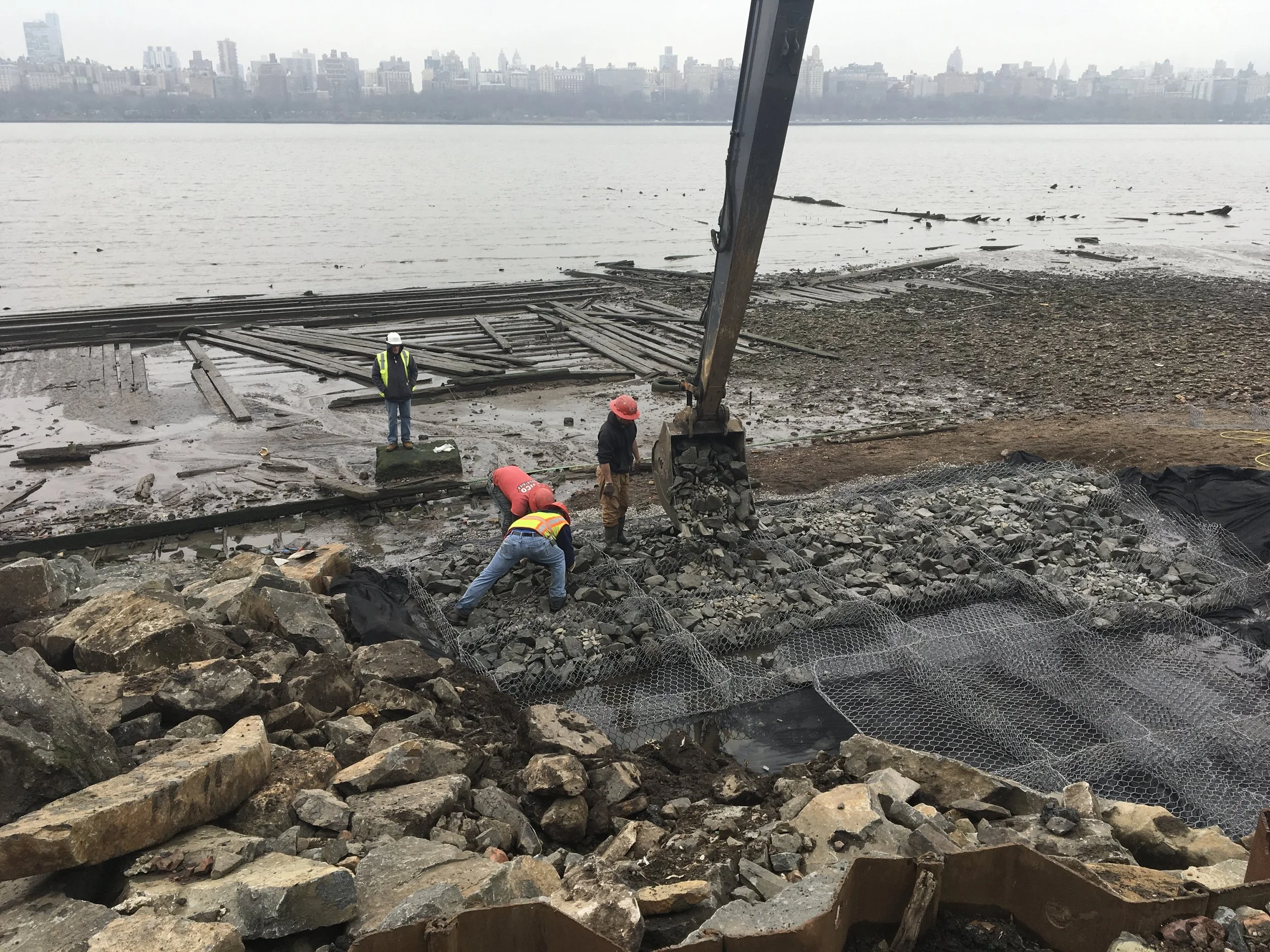 Construction workers wearing safety gear working on a damaged waterfront rail line, with a city skyline across the water in the background.