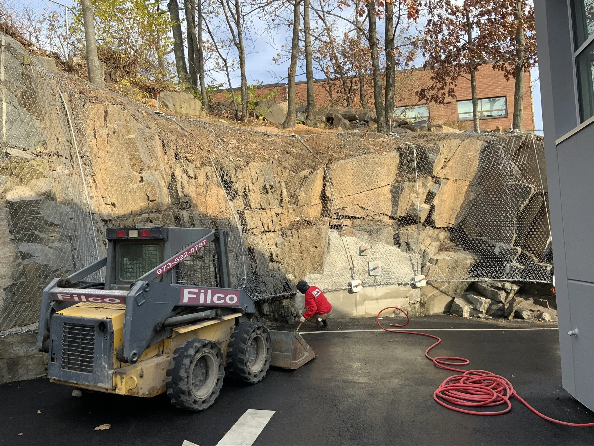 Construction worker in red shirt working on asphalt pavement at a rock face construction site with safety netting, a small bulldozer, and a coiled red hose.