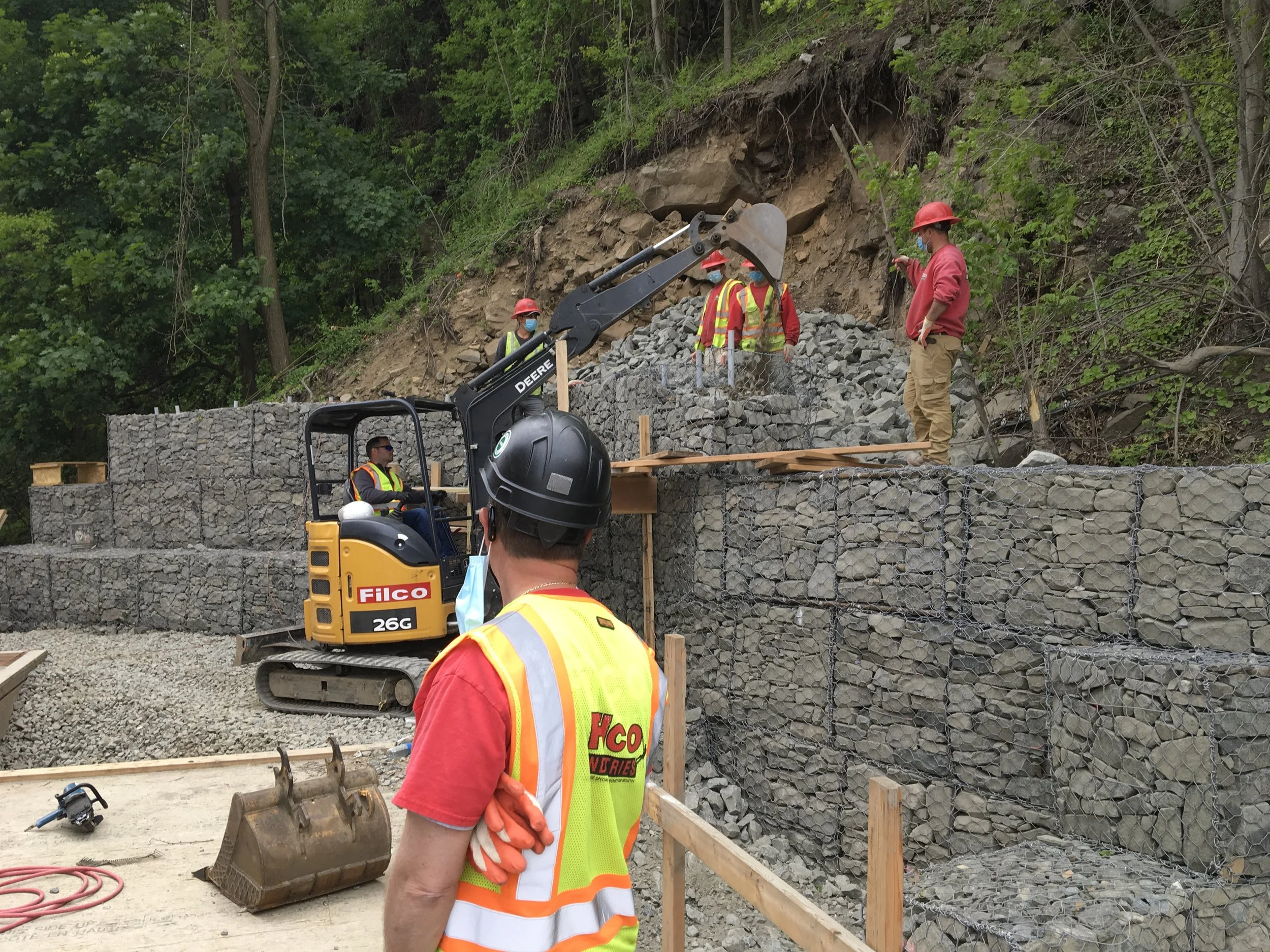 Construction workers building a rock retaining wall with gabion baskets on a hillside using a small excavator. The workers wear safety vests, helmets, and some have masks. There are trees and green foliage in the background.