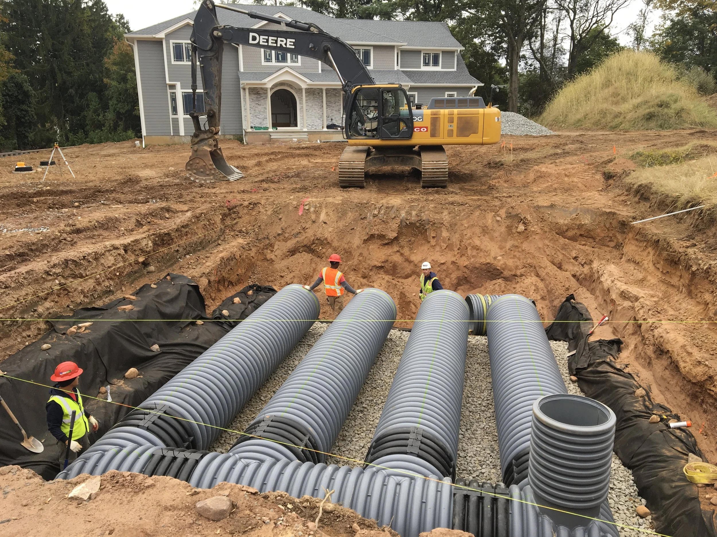 Construction site with large grey pipes being installed underground, workers in safety gear, an excavator on dirt, and a semi-completed house in the background.