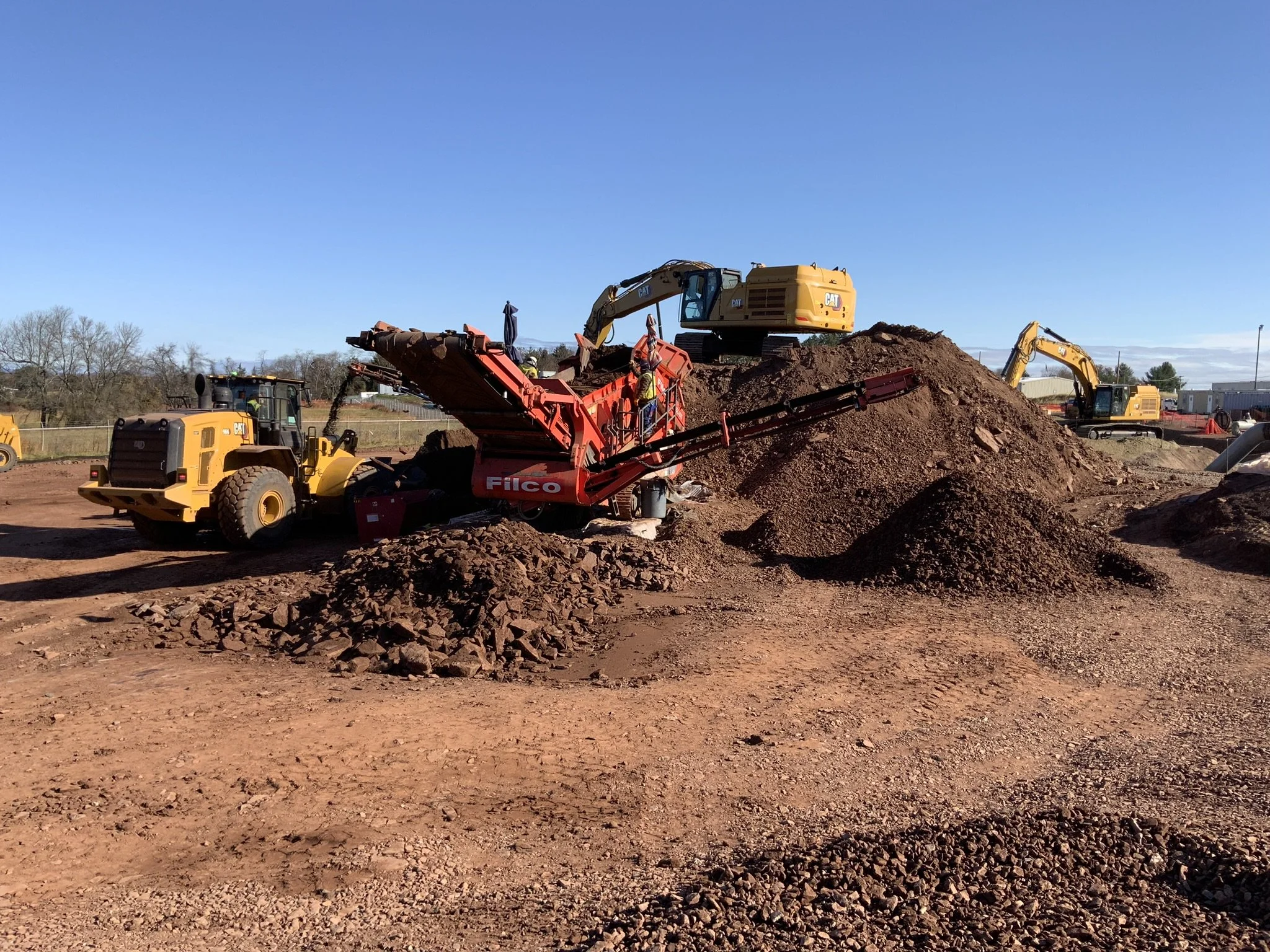 Construction site with dirt piles, bulldozers, and excavators working under a clear blue sky.