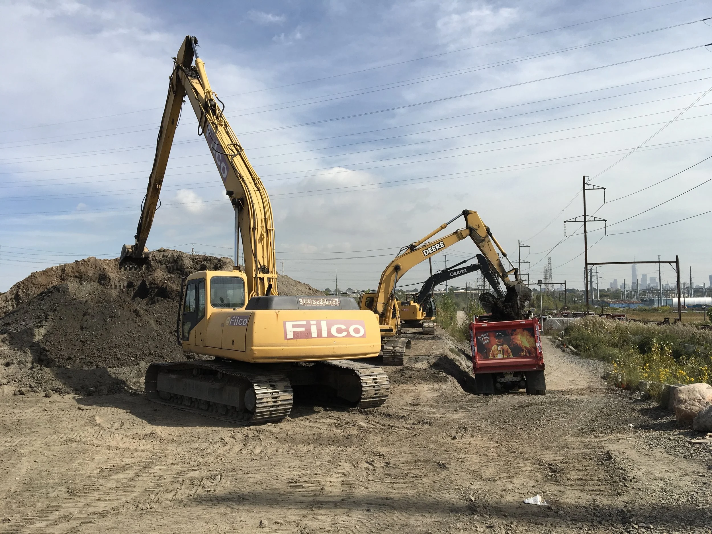 Construction site with yellow excavators loading dirt into a red dump truck, with power lines and a city skyline in the background.
