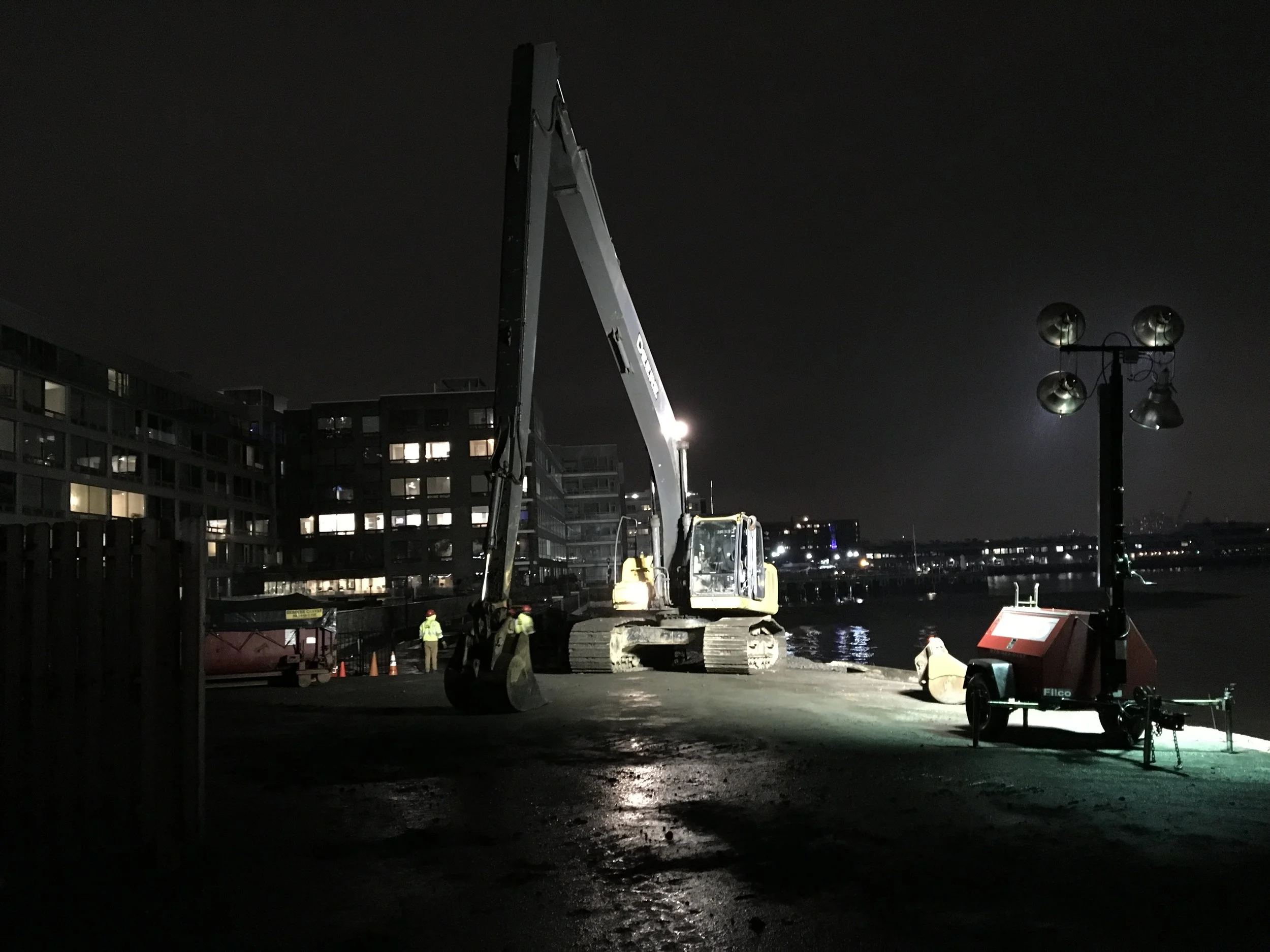 Nighttime construction scene by a waterway with a large hydraulic excavator, construction workers in safety gear, and portable lights