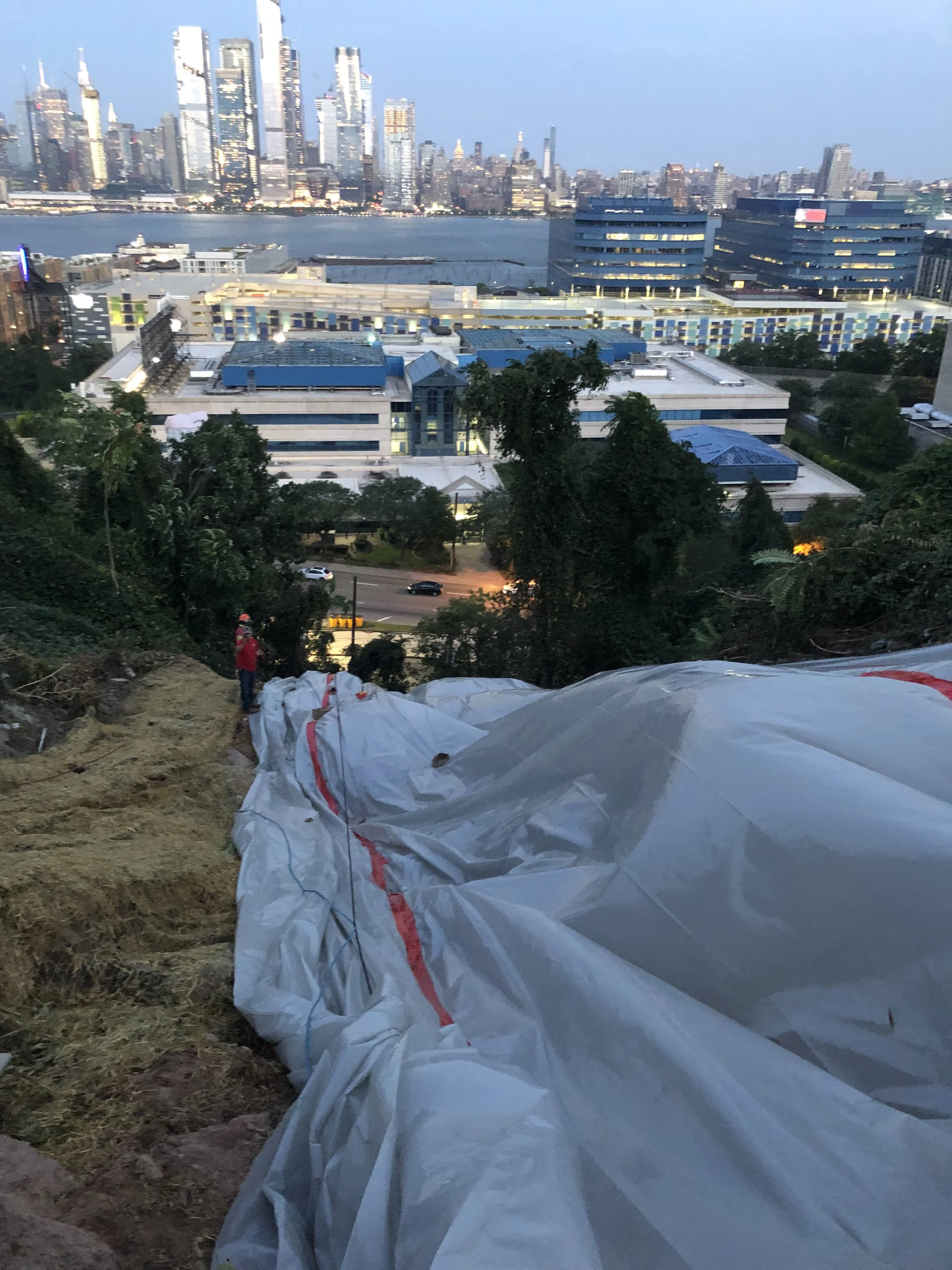 Construction site on a hillside with a large striped tarp or plastic sheeting, overlooking a city skyline and river at dusk.