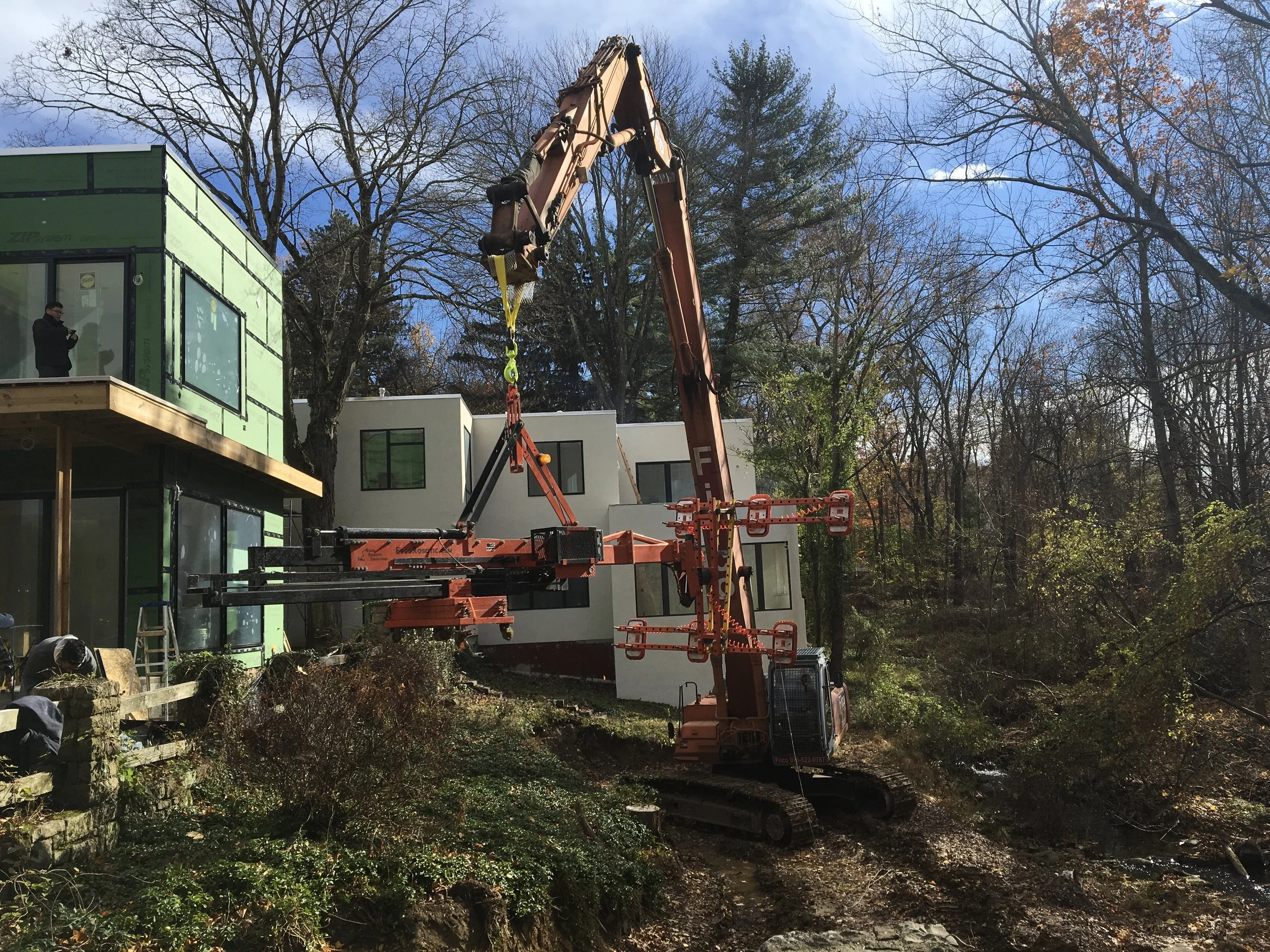A construction crane lifting materials near a modern house under construction, surrounded by trees with orange and bare branches, under a blue sky with some clouds.