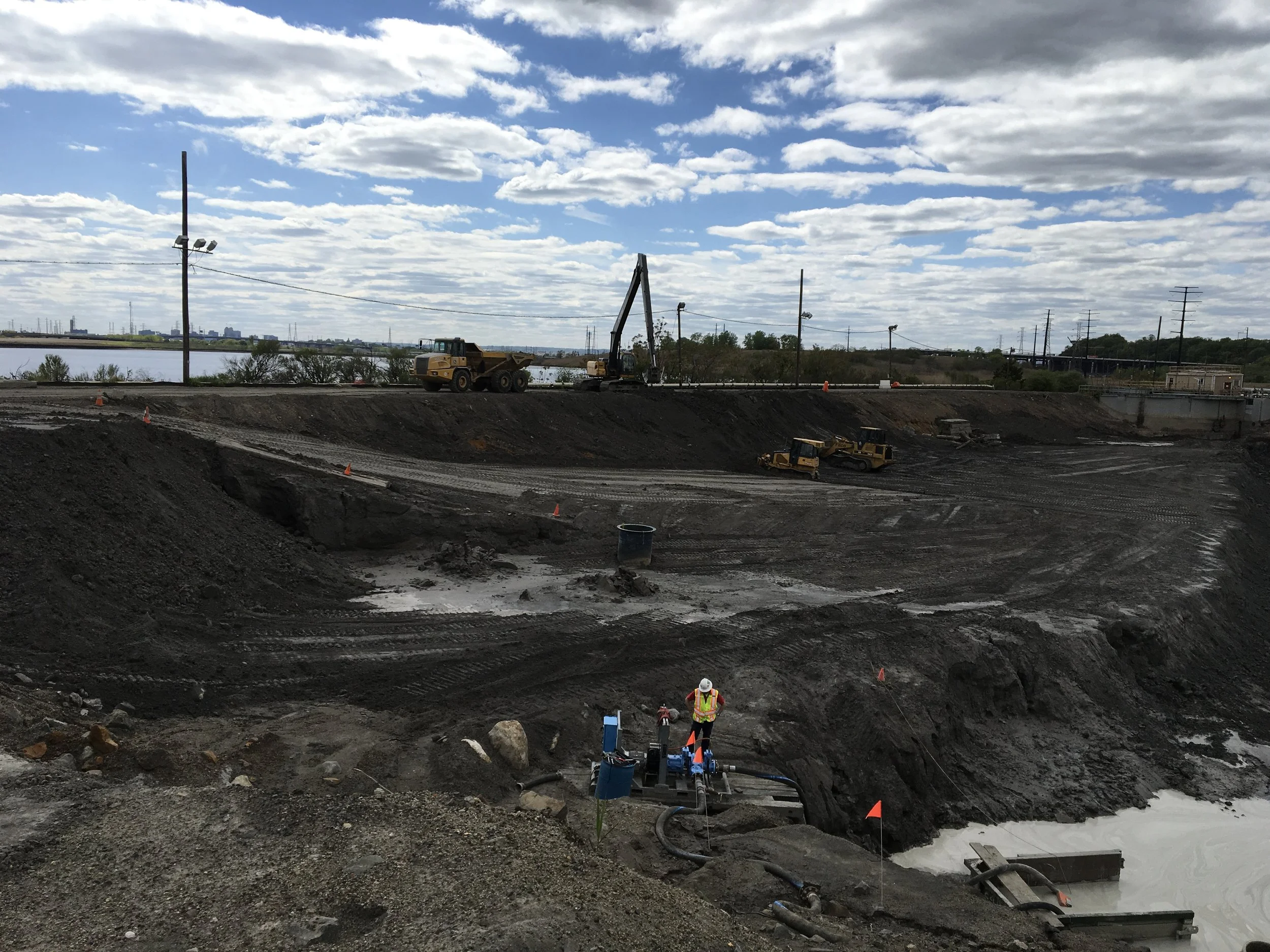Construction site with heavy machinery and a worker in a safety vest and helmet near a muddy area with orange flags, under a partly cloudy sky.