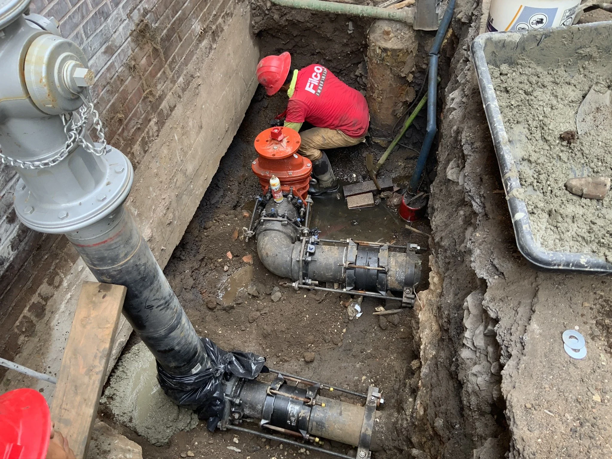 A construction worker wearing a red hard hat and red shirt working in a deep excavated trench installing or repairing large water pipes with valves.
