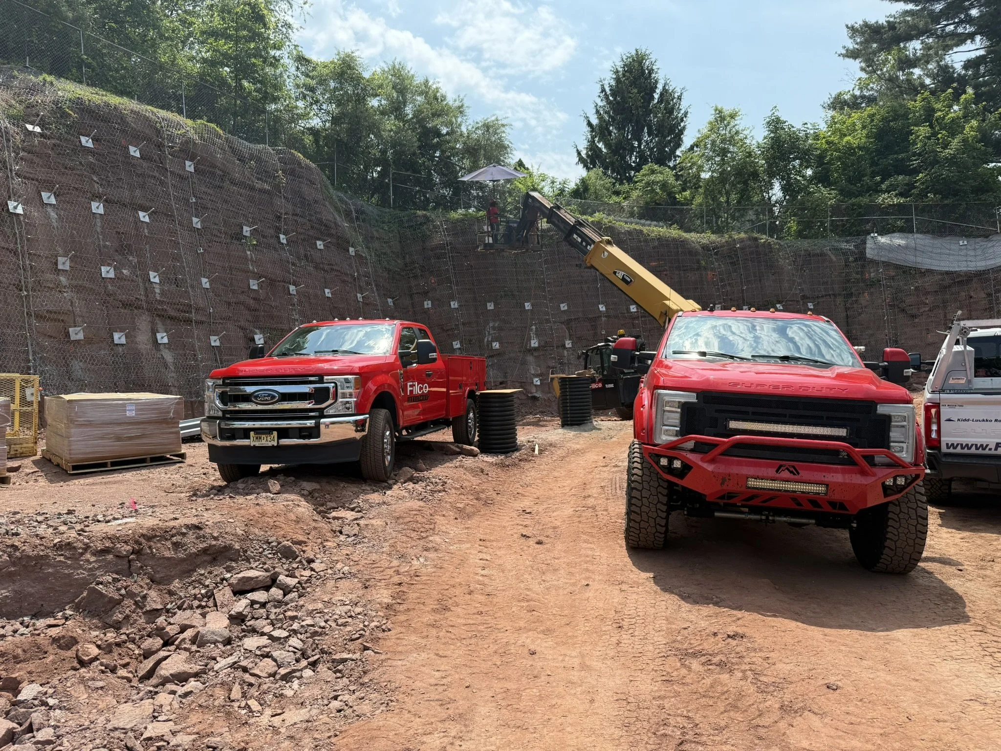 Construction site with red trucks, a crane, and a rocky dirt path, with a hill in the background.