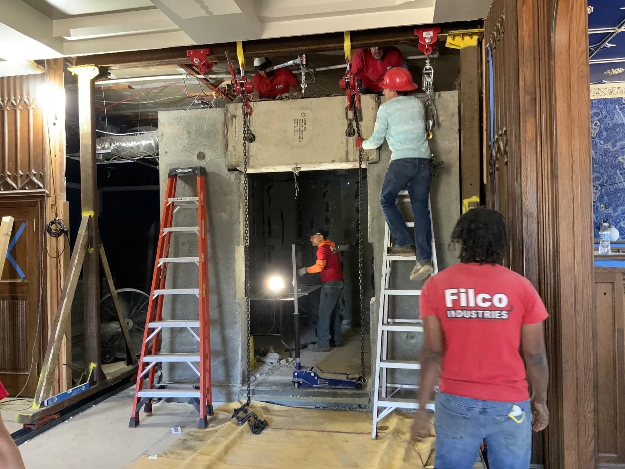 Construction workers installing or repairing a structure inside a building. Several workers wearing safety helmets and red shirts, some on ladders and others on the ground, are working with tools and chains. Construction equipment, ladders, and open 