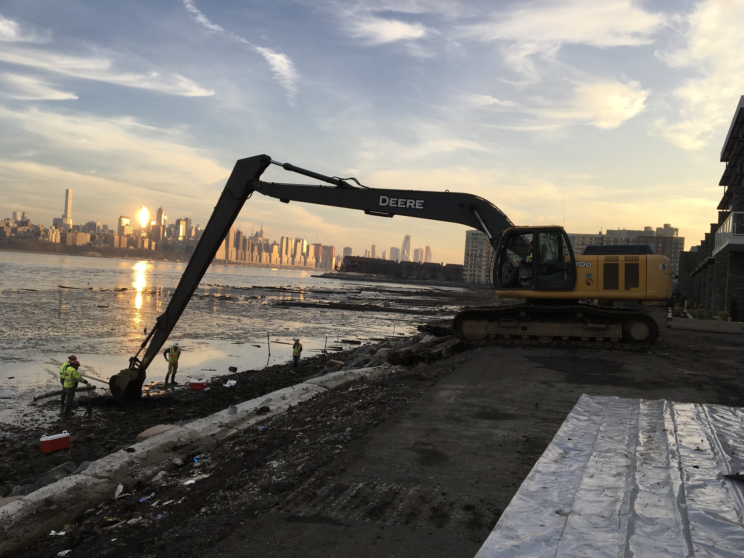 Construction workers working by the water with a large excavator and a city skyline in the background at sunset.