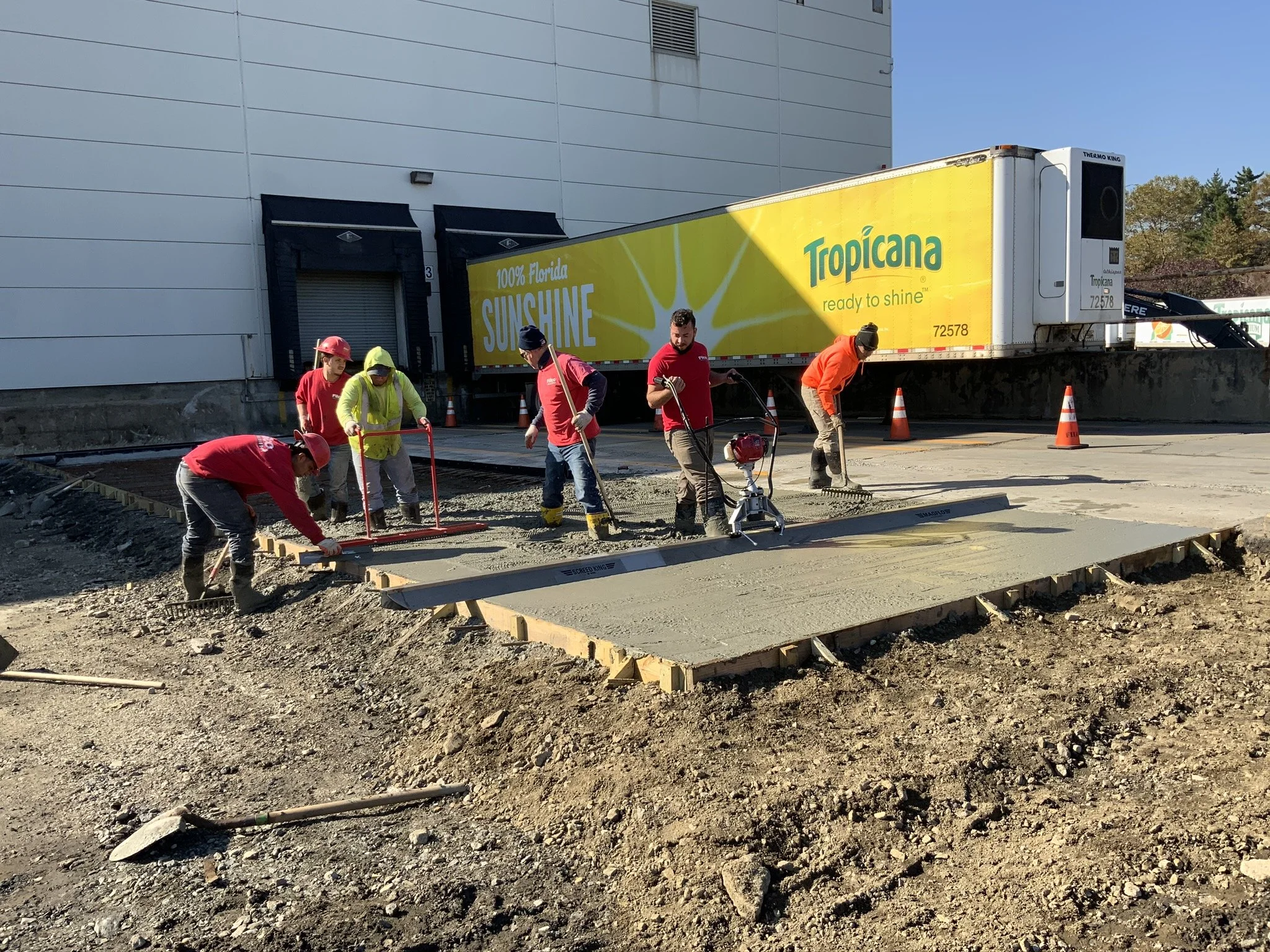 Construction workers pouring concrete for a sidewalk, with a Tropicana truck parked in the background.