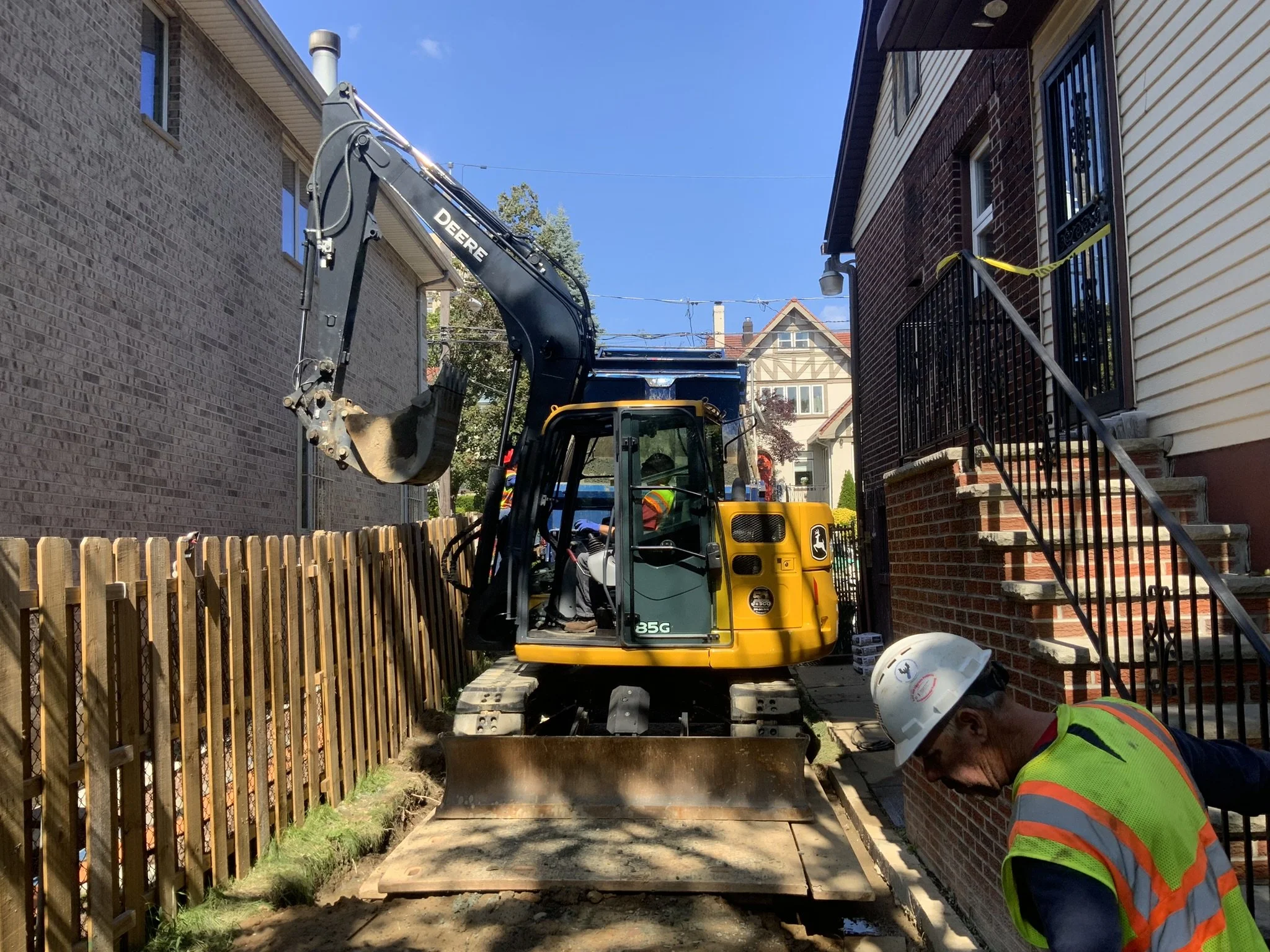 A construction worker in a safety vest and helmet is working near a house with stairs, while a small John Deere excavator is digging a trench between the houses.