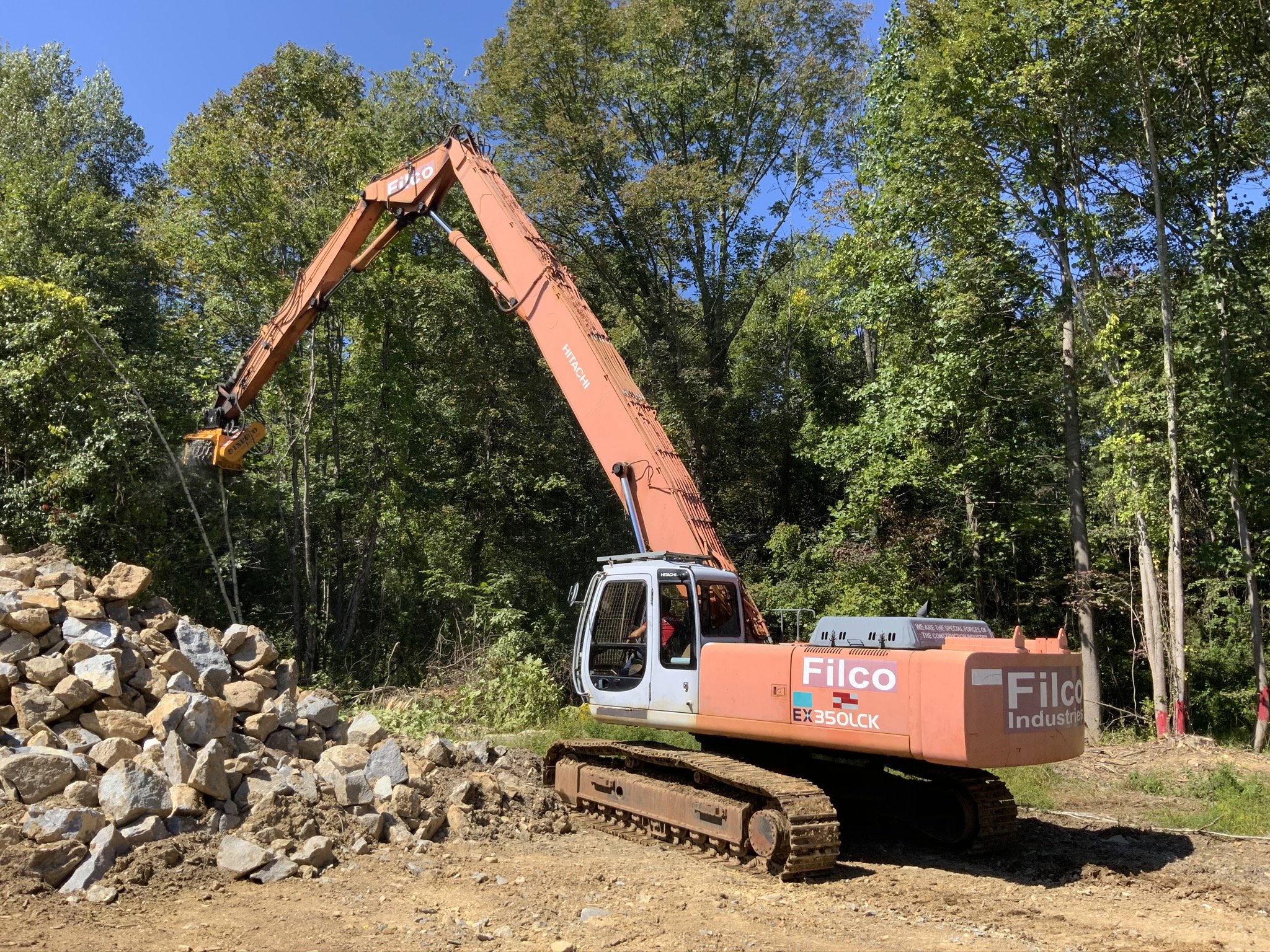 A pink excavator with the brand name 'Filco' and model 'EX350LCK' on the side, working on a construction site surrounded by green trees, lifting a bucket full of rocks.