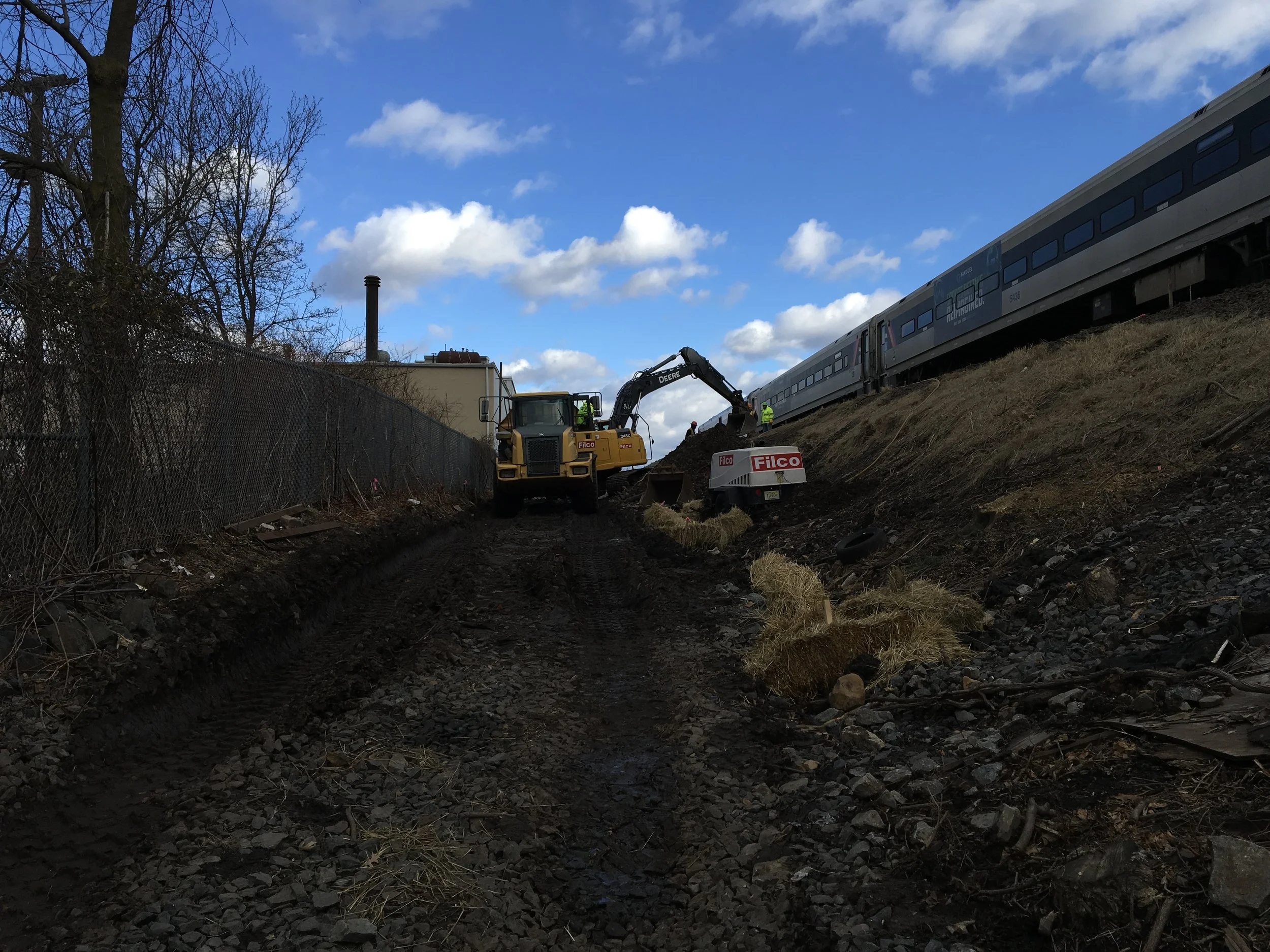 Construction work on a railway track with a yellow backhoe and workers, on a dirt path beside a chain-link fence, with a train and blue sky with clouds in the background.