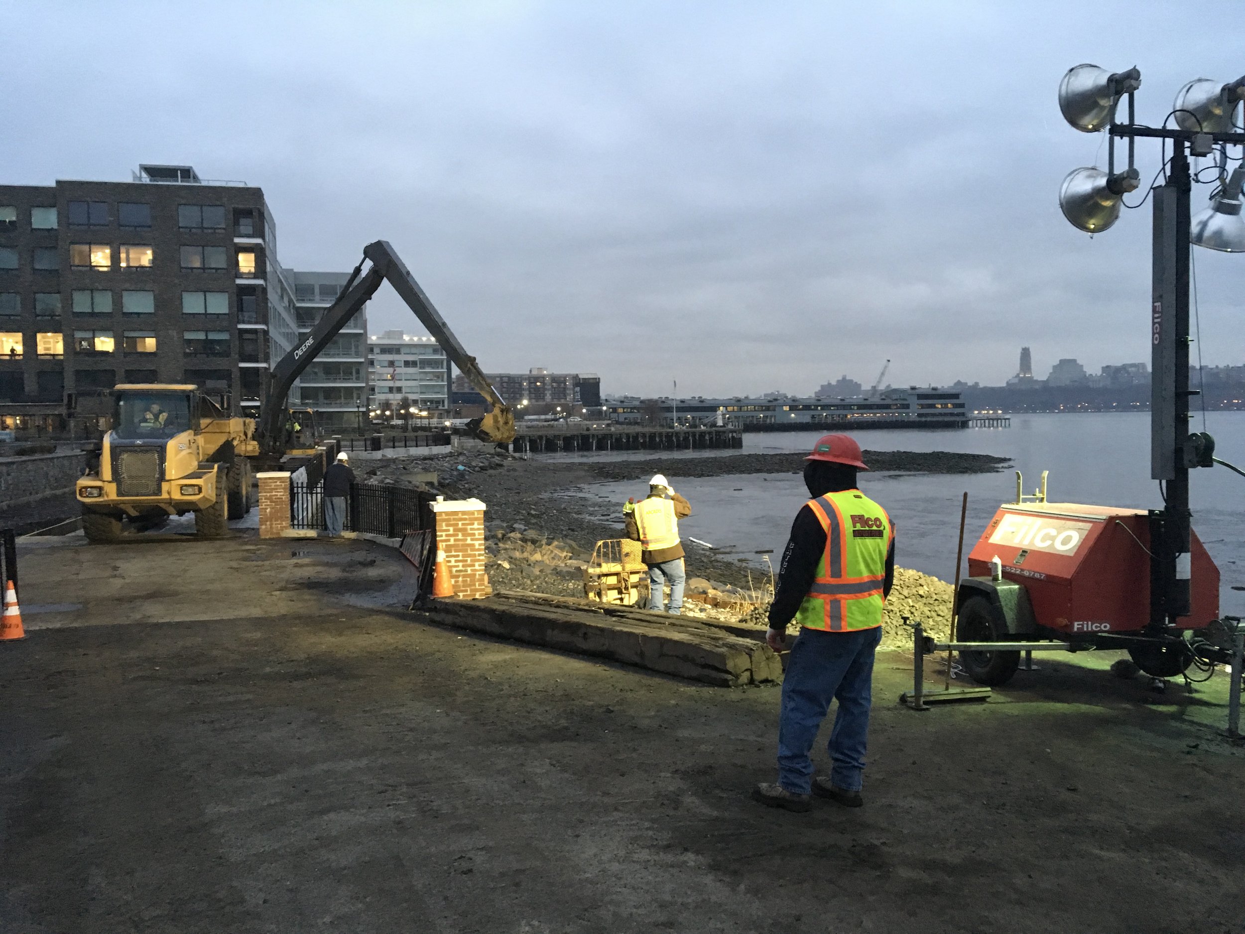 Construction workers and equipment at a waterfront site during dusk, with buildings and a pier in the background.