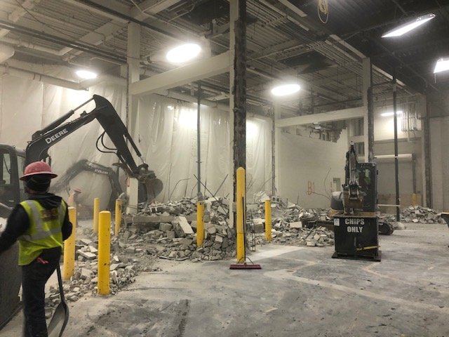 Construction workers and machinery demolishing a wall inside a building, with debris on the ground and yellow safety posts.