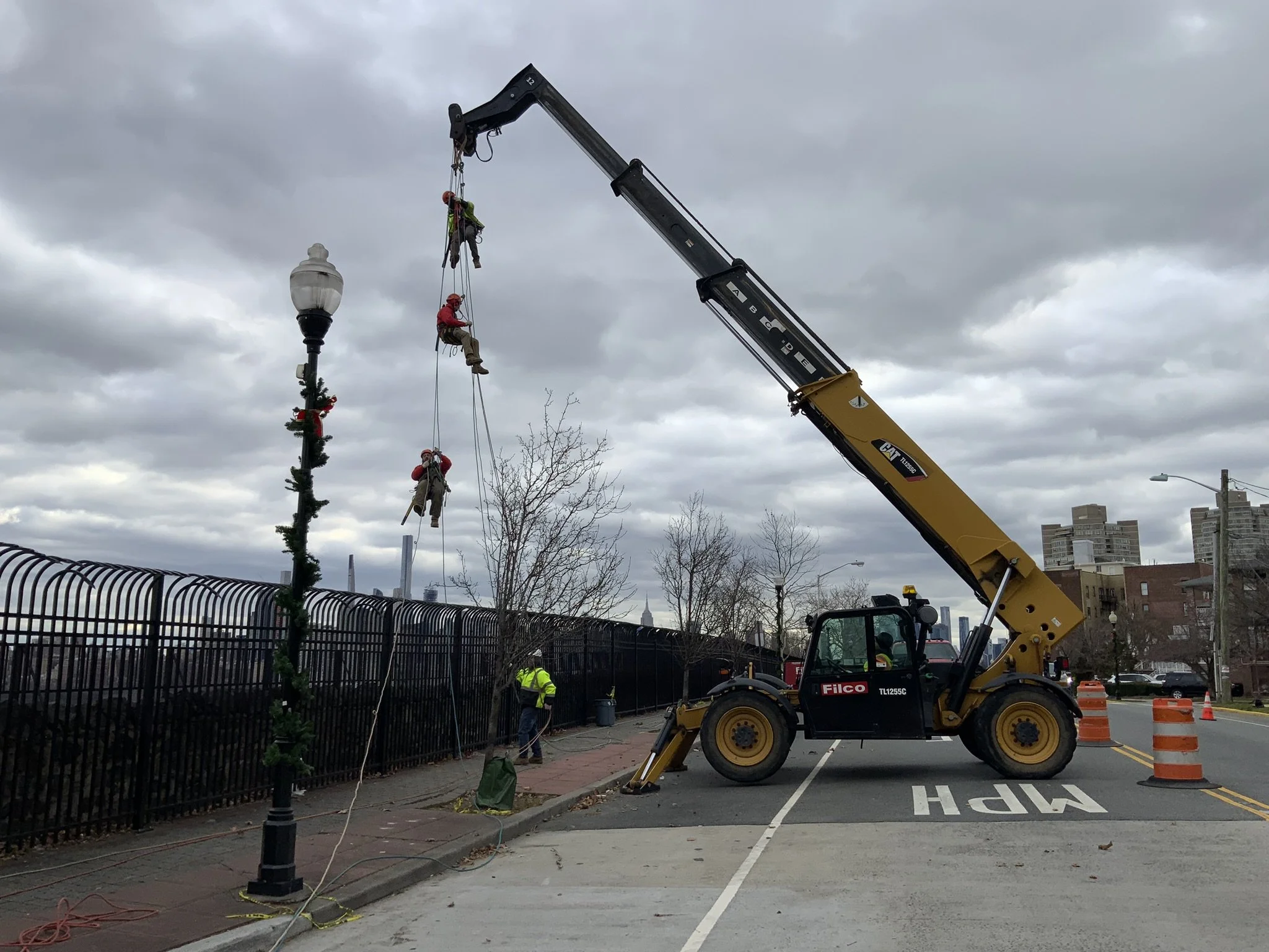 A crane lifting three workers dressed in red and khaki, hanging from ropes, near a holiday-decorated lamppost along a city sidewalk, with workers and orange traffic barrels nearby.