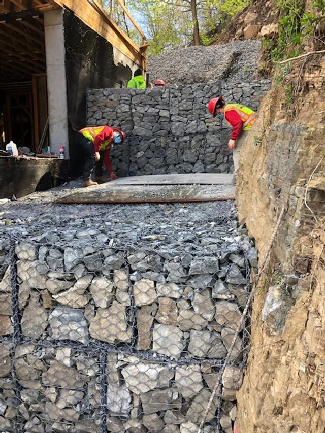 Two construction workers wearing safety helmets and vests work on a stone retaining wall on a hillside, with a collapsed section of stone and wire mesh in the foreground.