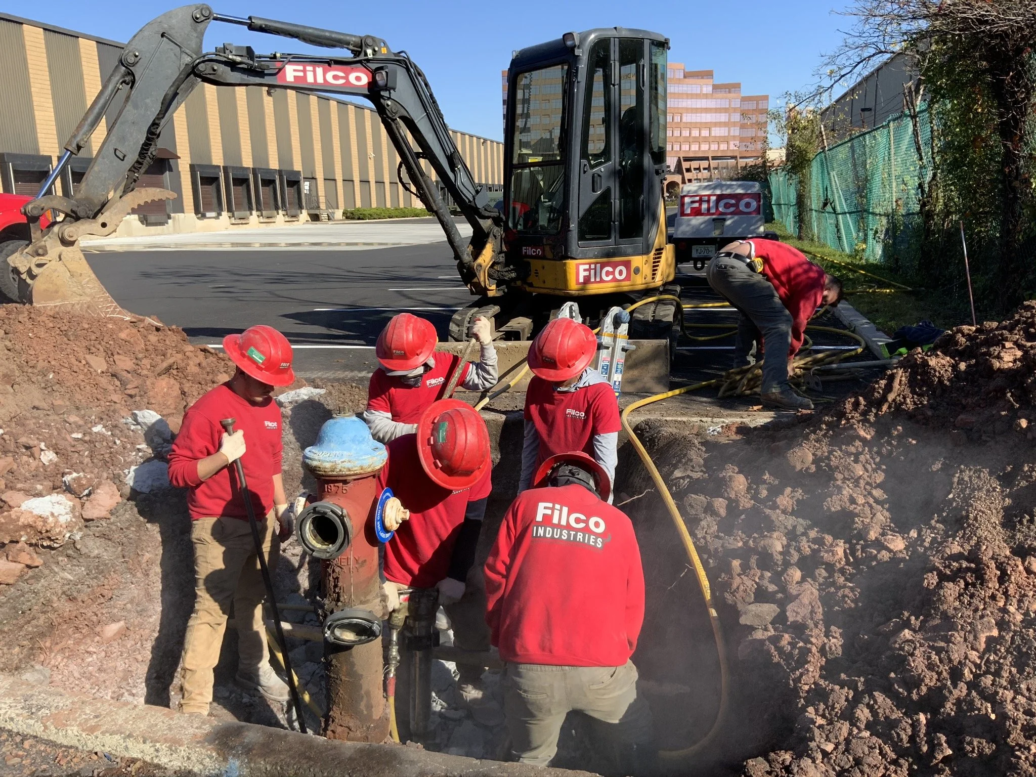Workers in red uniforms and helmets repairing or installing underground pipes using equipment and a small excavator at a construction site near a parking lot.
