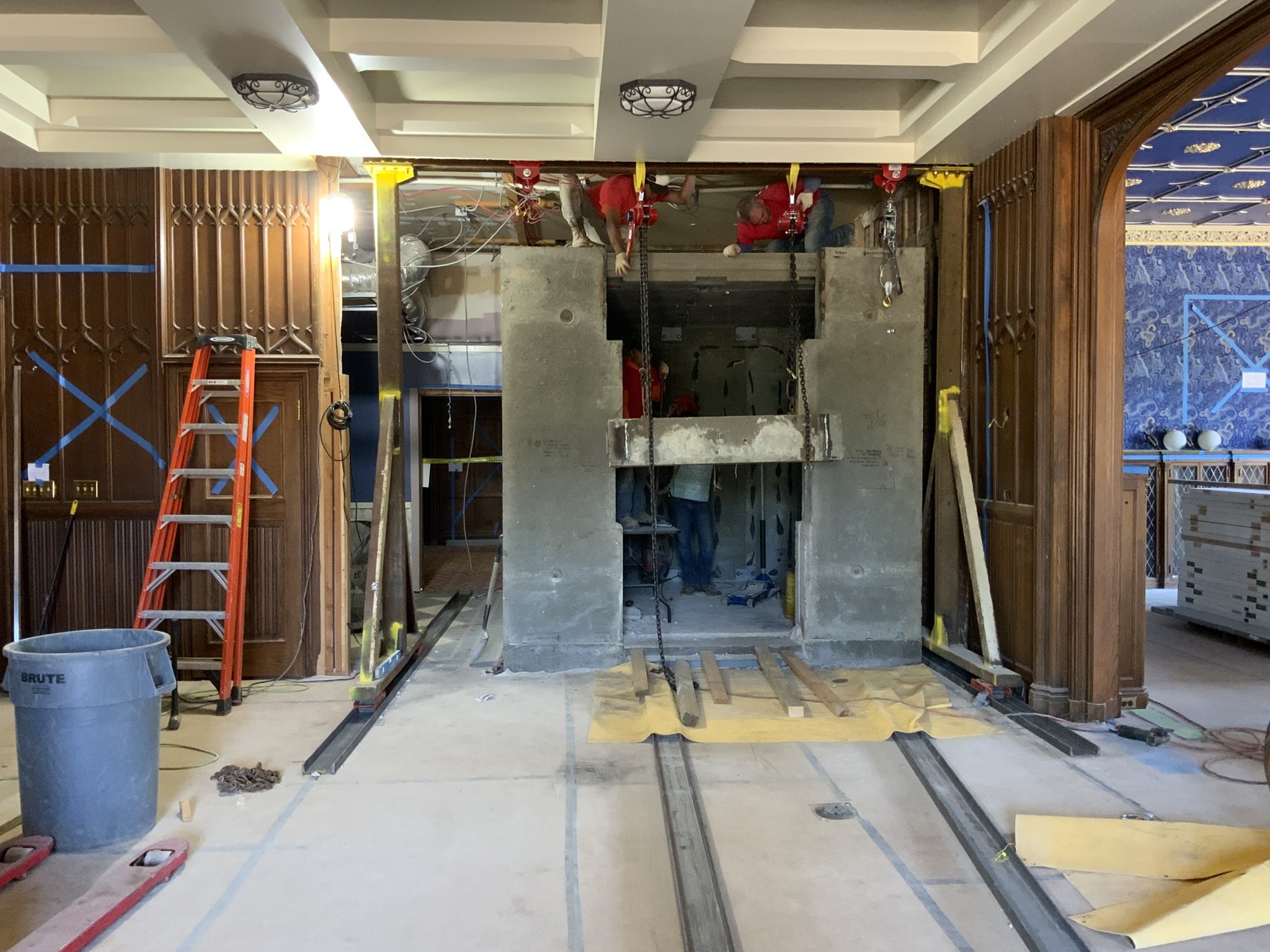Construction workers install an elevator shaft in a building interior with ornate wood wall paneling and decorative ceiling.