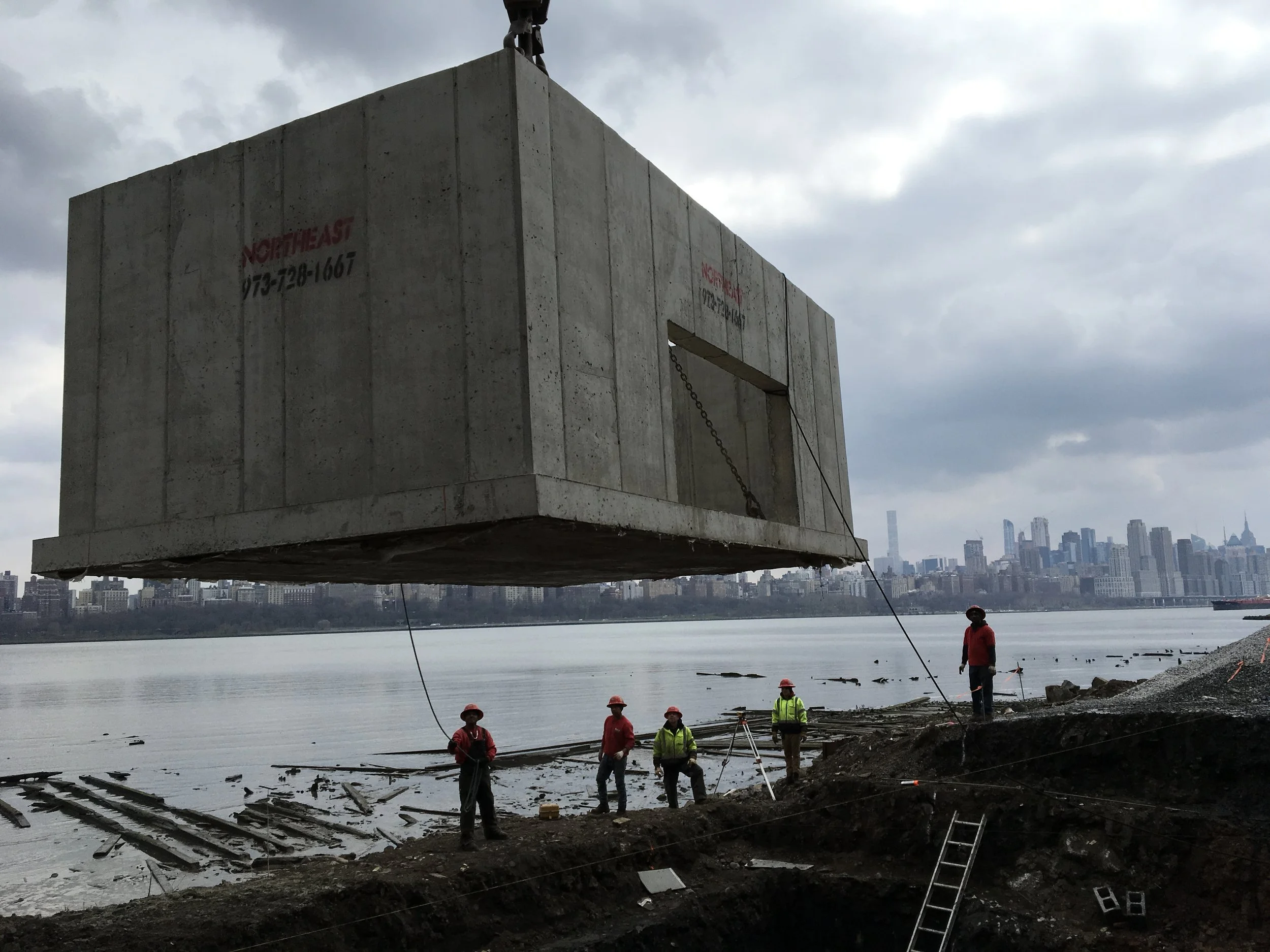 Construction workers in safety gear and hard hats work near a river, with a large concrete structure being lifted by a crane, city skyline in the background.