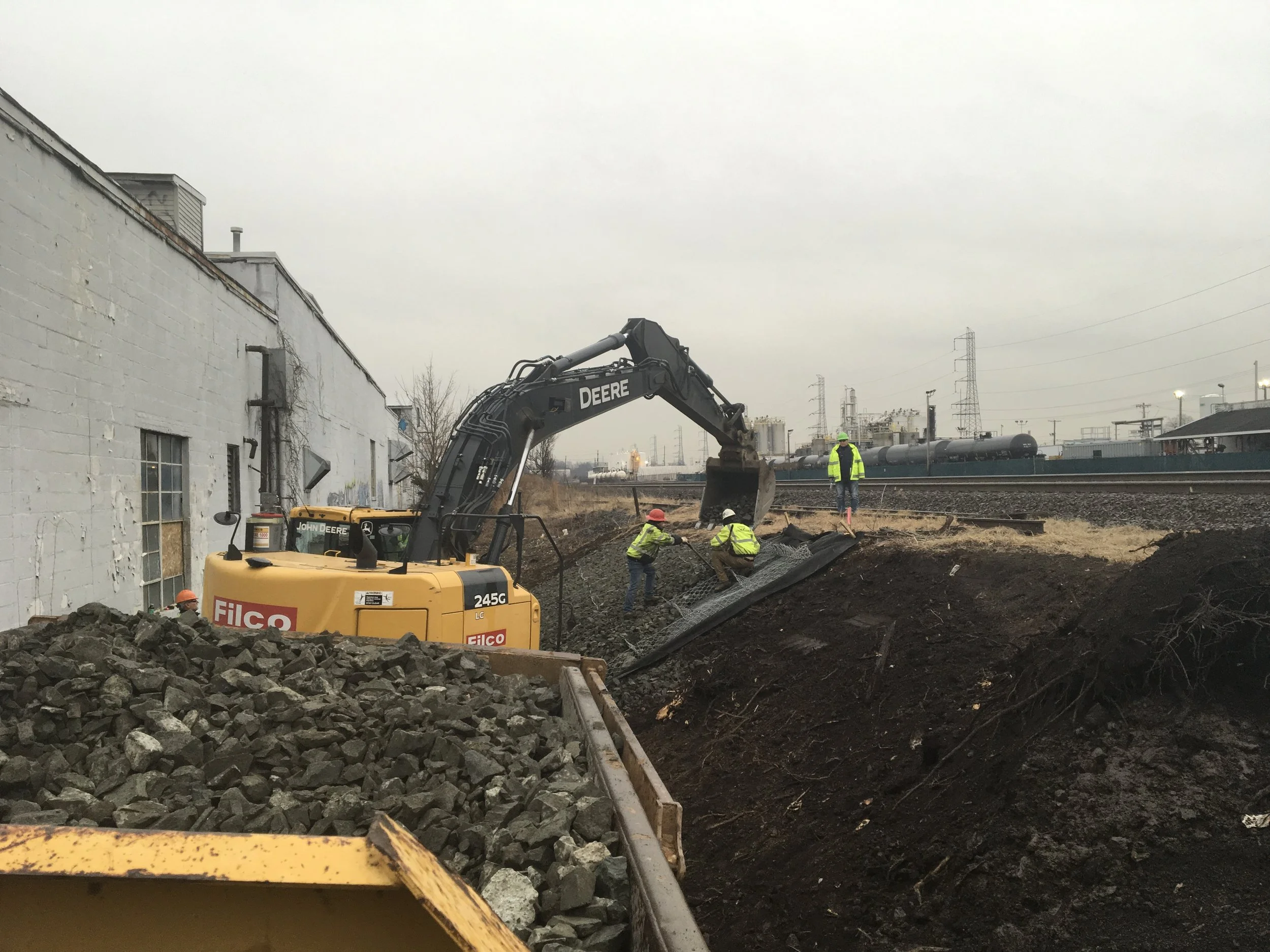 Construction workers installing a barrier next to a railroad track with industrial buildings and power lines in the background.