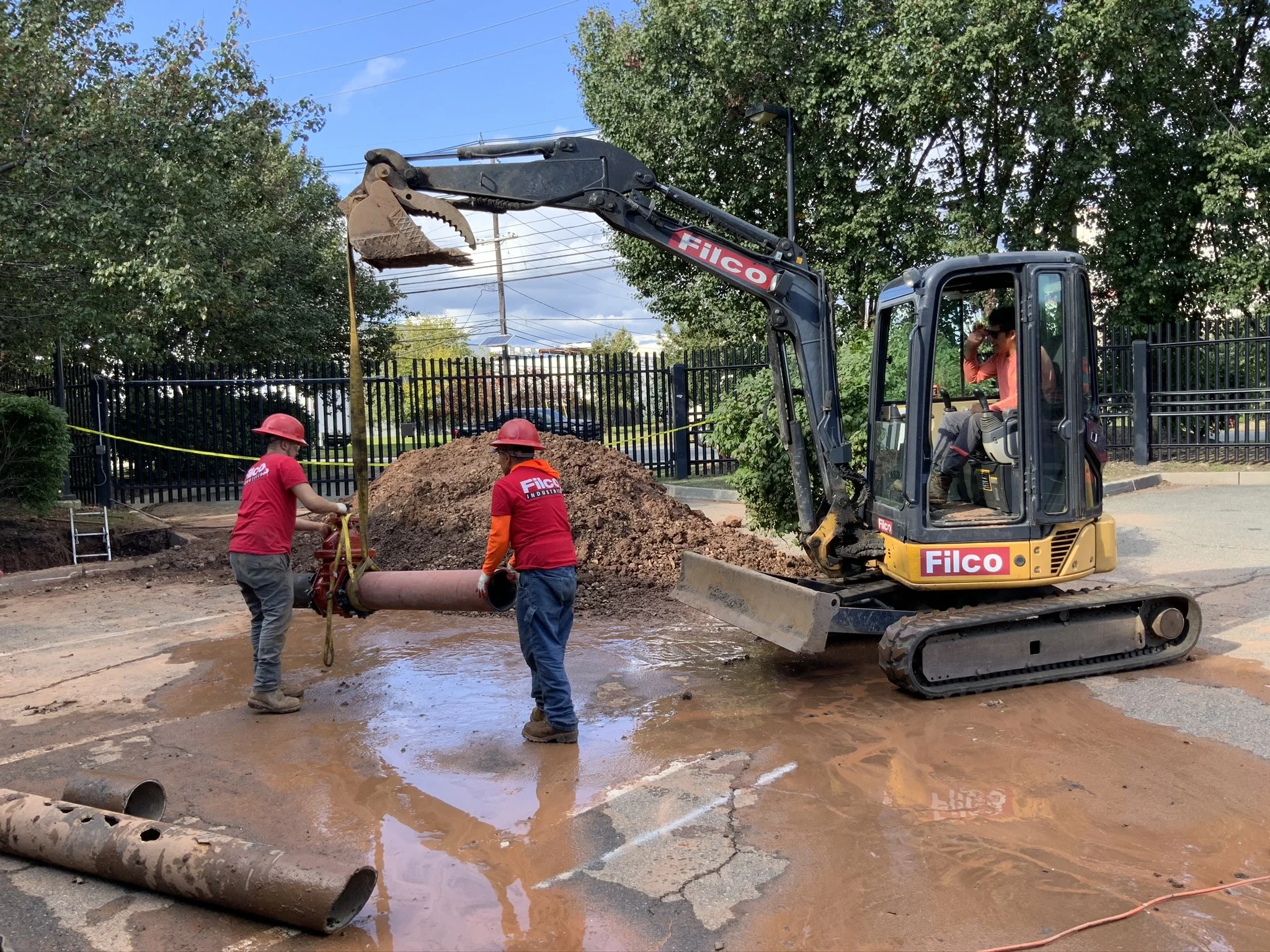 Workers in red shirts and helmets using a mini excavator to lift a pipe during a construction project.