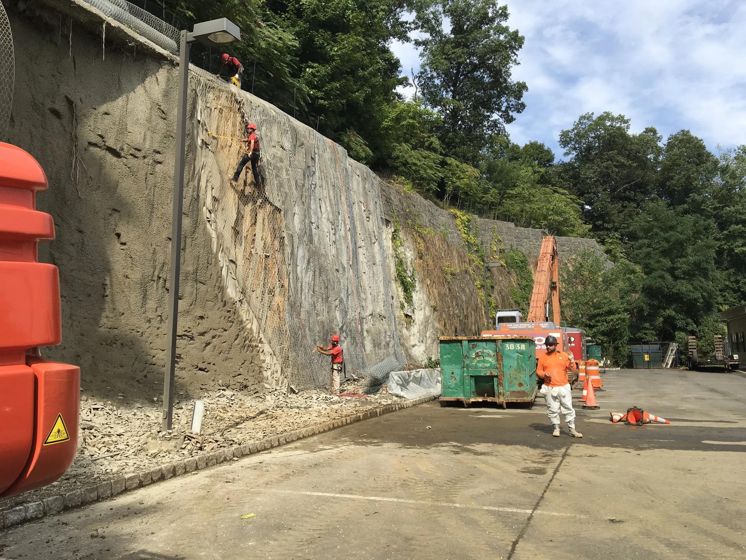 Construction workers wearing safety gear working on a rock face, with some on the ground and others climbing on the rock. Construction equipment, cones, and a large excavator are present in a parking lot area, with trees and a blue sky in the backgro