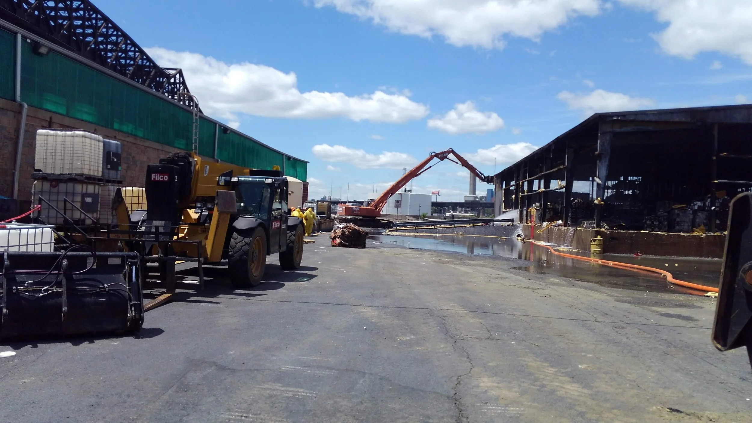 Industrial area with construction and cleaning equipment, including a large excavator and a loader, next to a structure with burnt and damaged walls, and water on the ground under a partly cloudy sky.