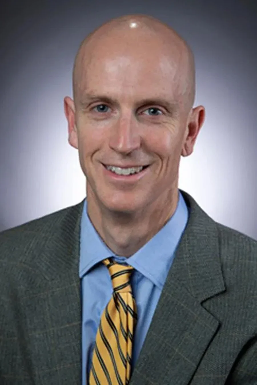 A bald man wearing a gray suit, blue shirt, and yellow striped tie, smiling in front of a gray background.