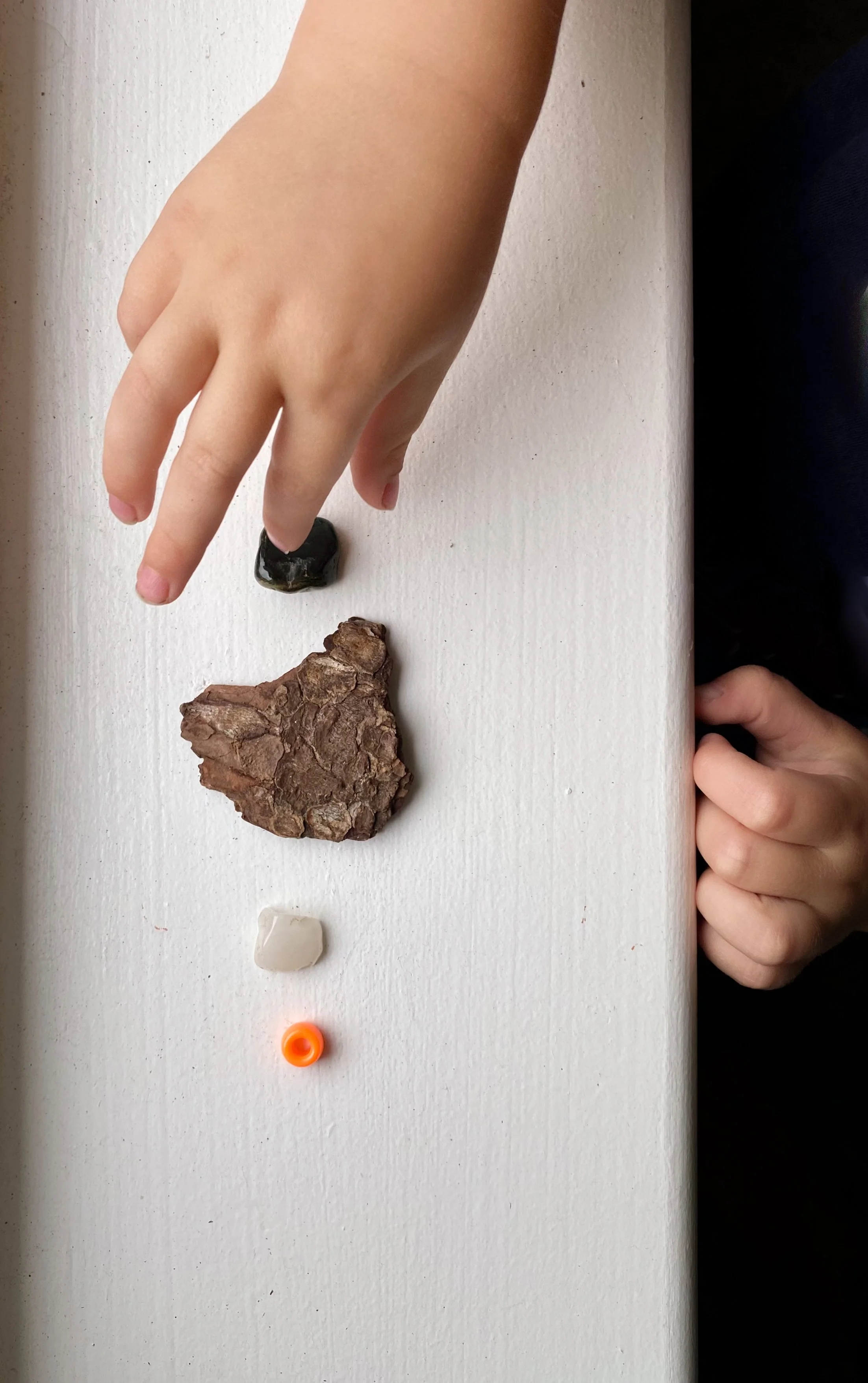 Children's hands arranging stones and a small orange bead on a white surface.