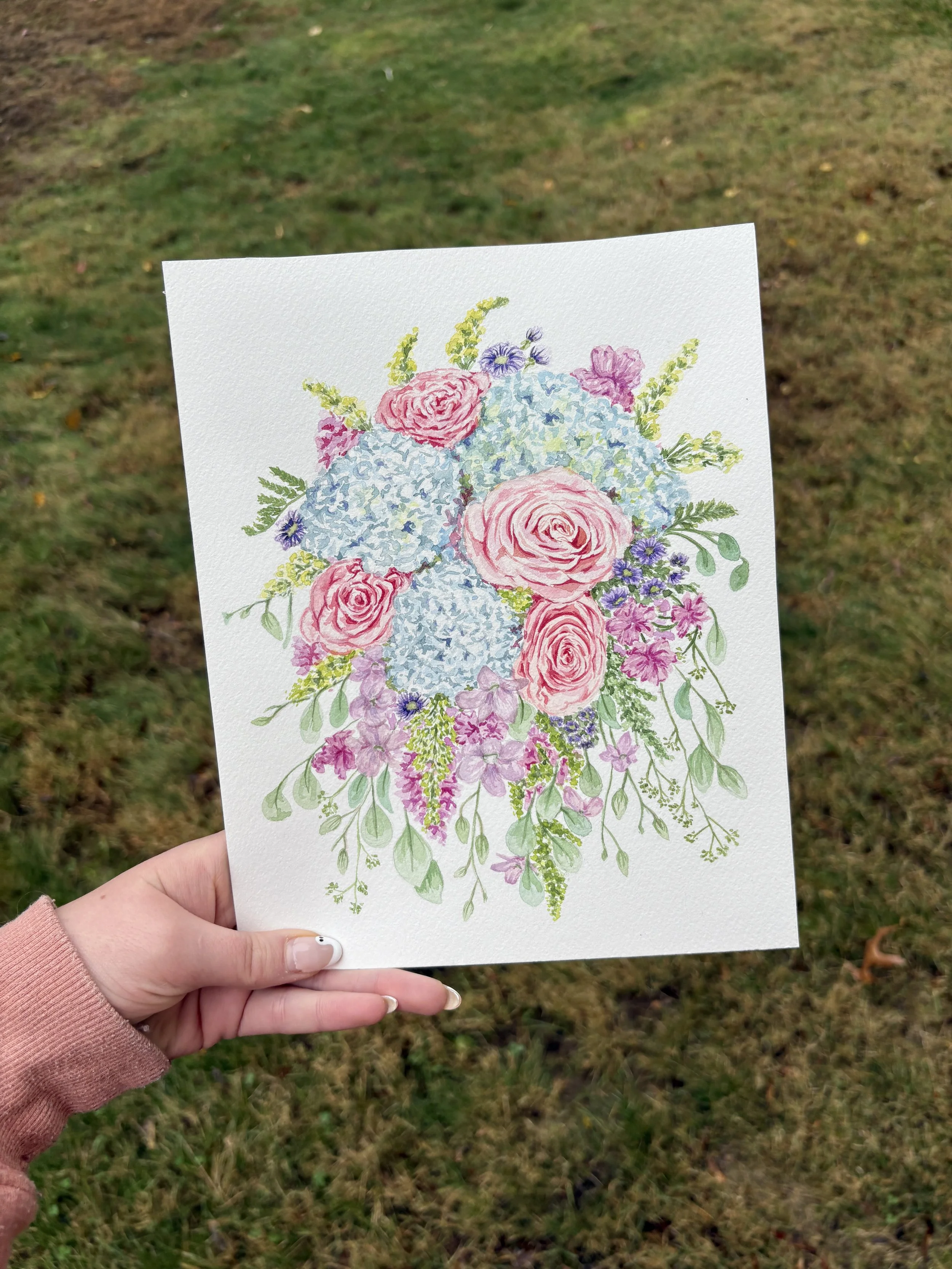 A hand holding a watercolor floral painting of pink roses, blue hydrangeas, and various smaller flowers against a grassy background.