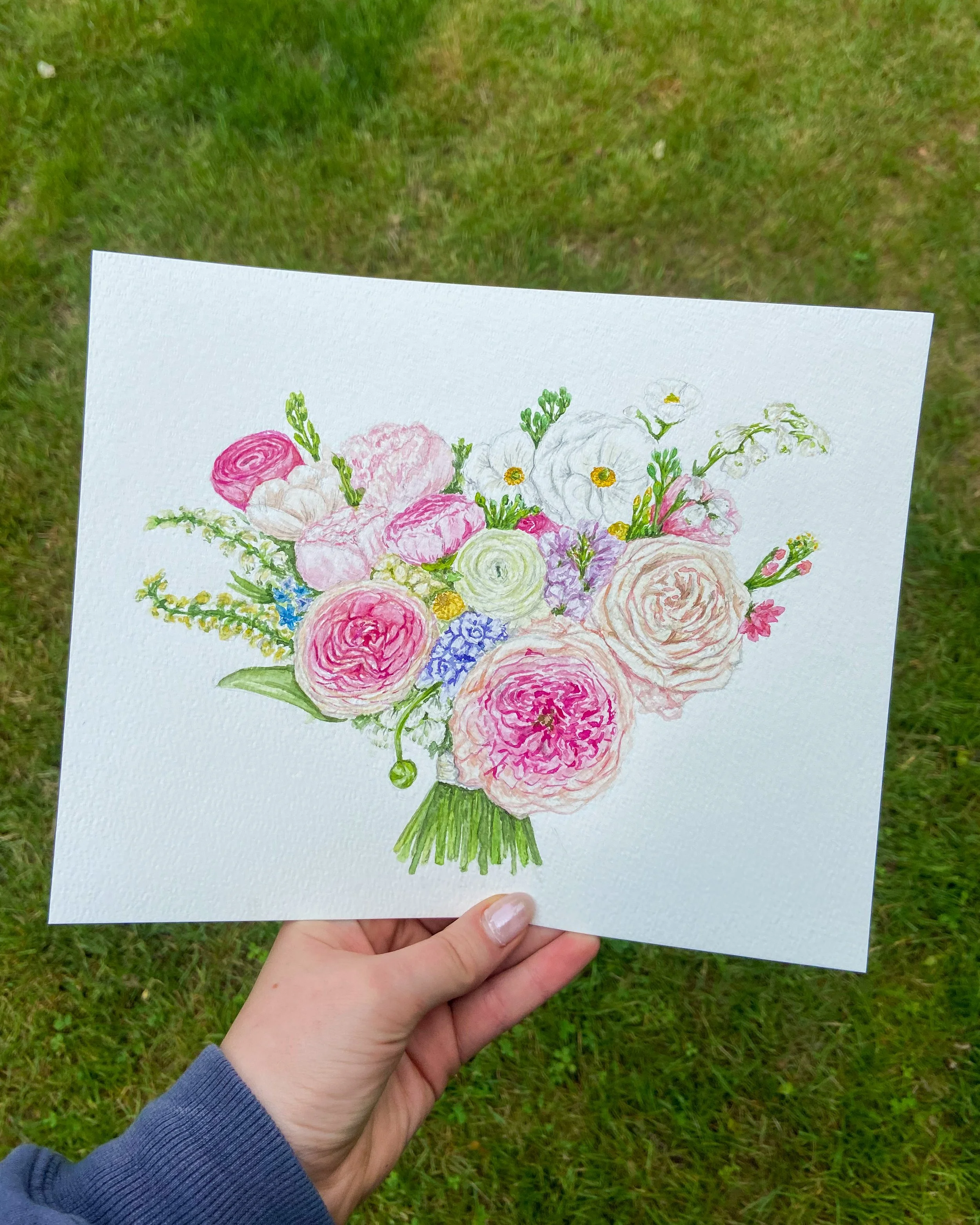 A hand holding a watercolor painting of a bouquet of pink, white, purple, and blue flowers against a grassy background.