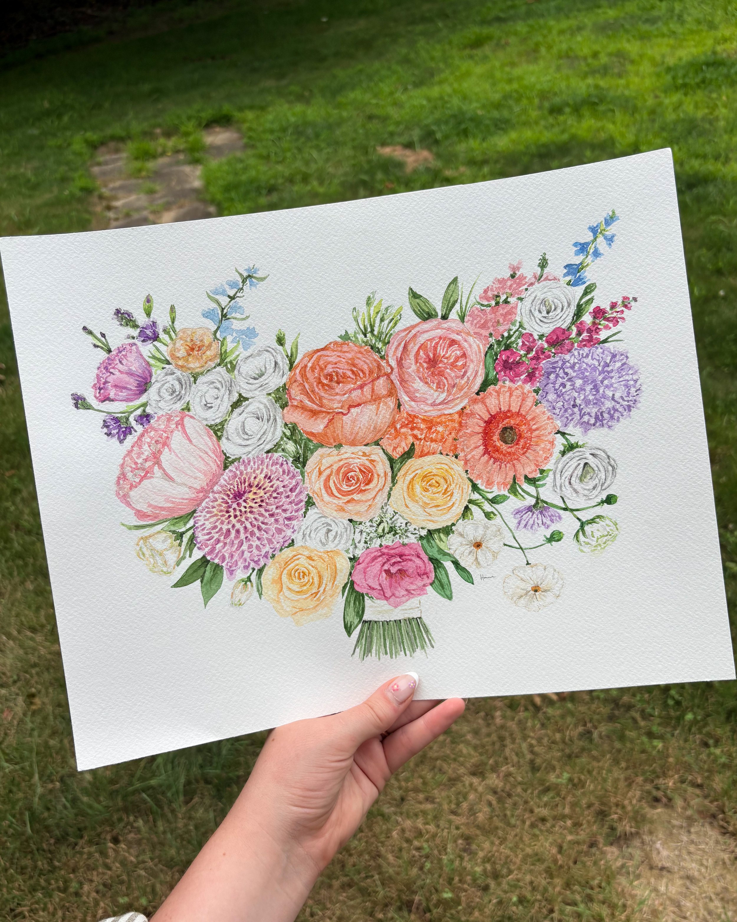 Hand holding a watercolor painting of a mixed bouquet of flowers outdoors with grass and stones in the background.