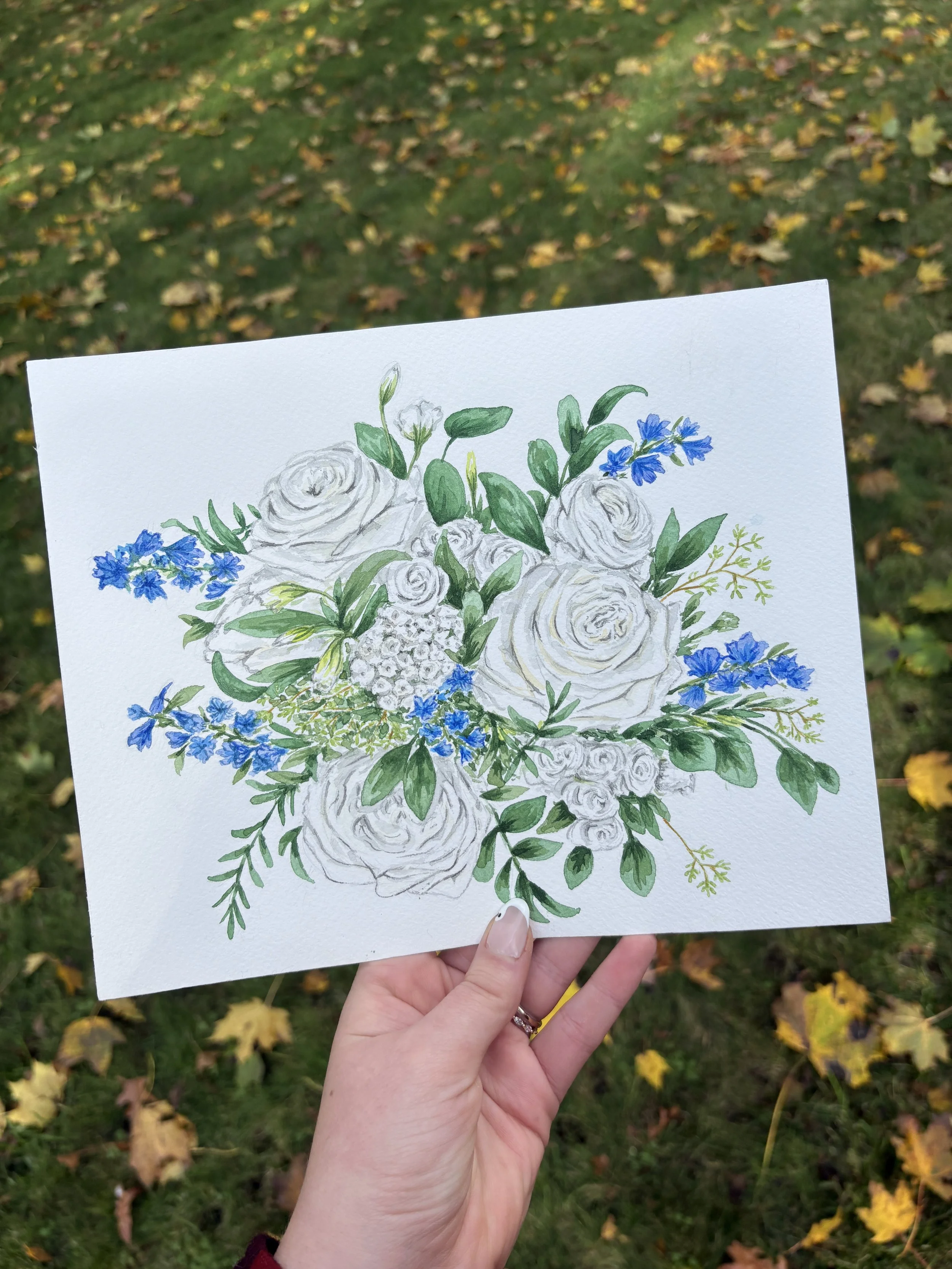 A person holding a watercolor painting of white roses and blue flowers against a background of fall leaves on the ground.