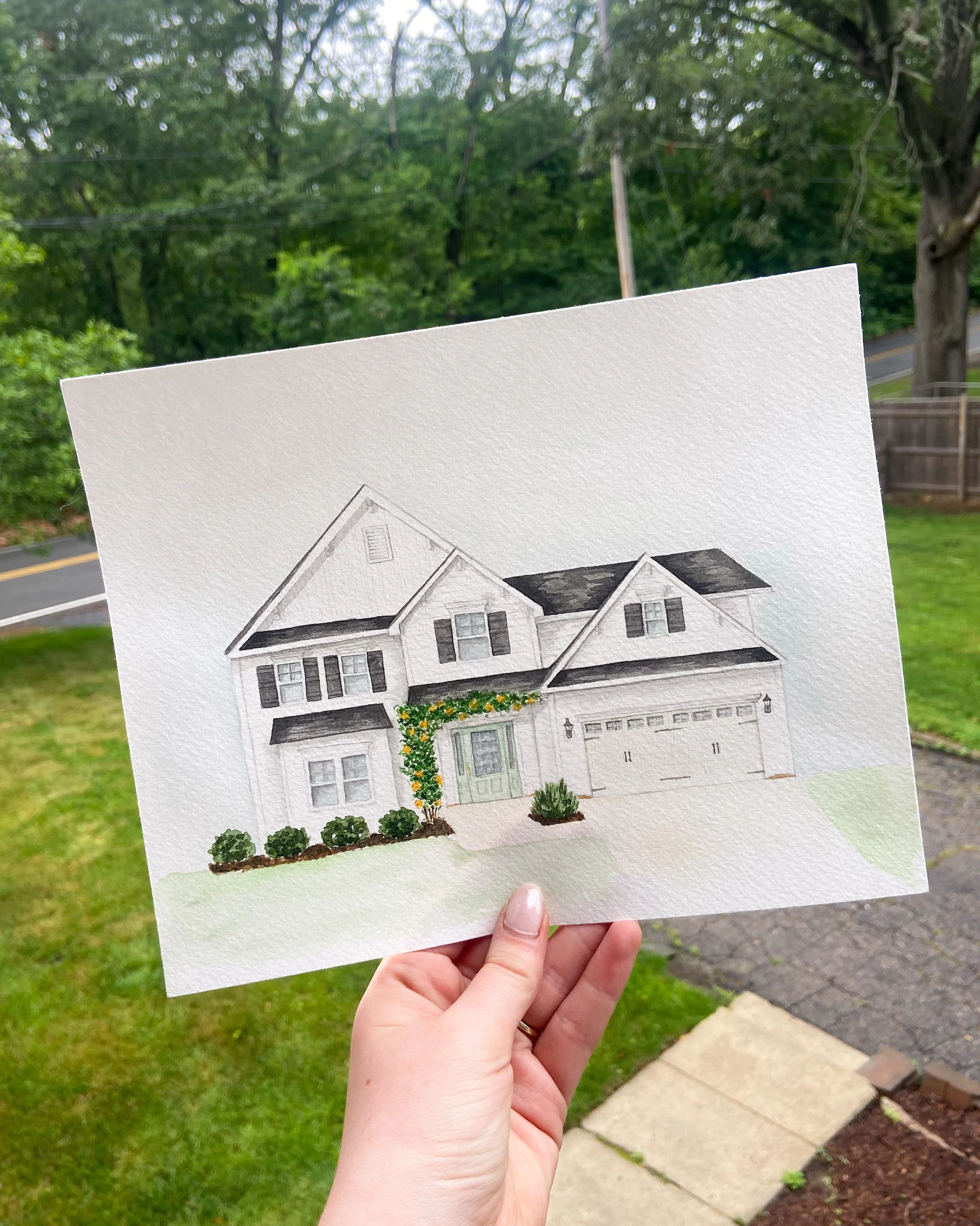 Hand holding a watercolor painting of a house in a backyard larger than the actual house, with a white exterior, black shutters, and an attached garage, against a backdrop of green trees and a street.