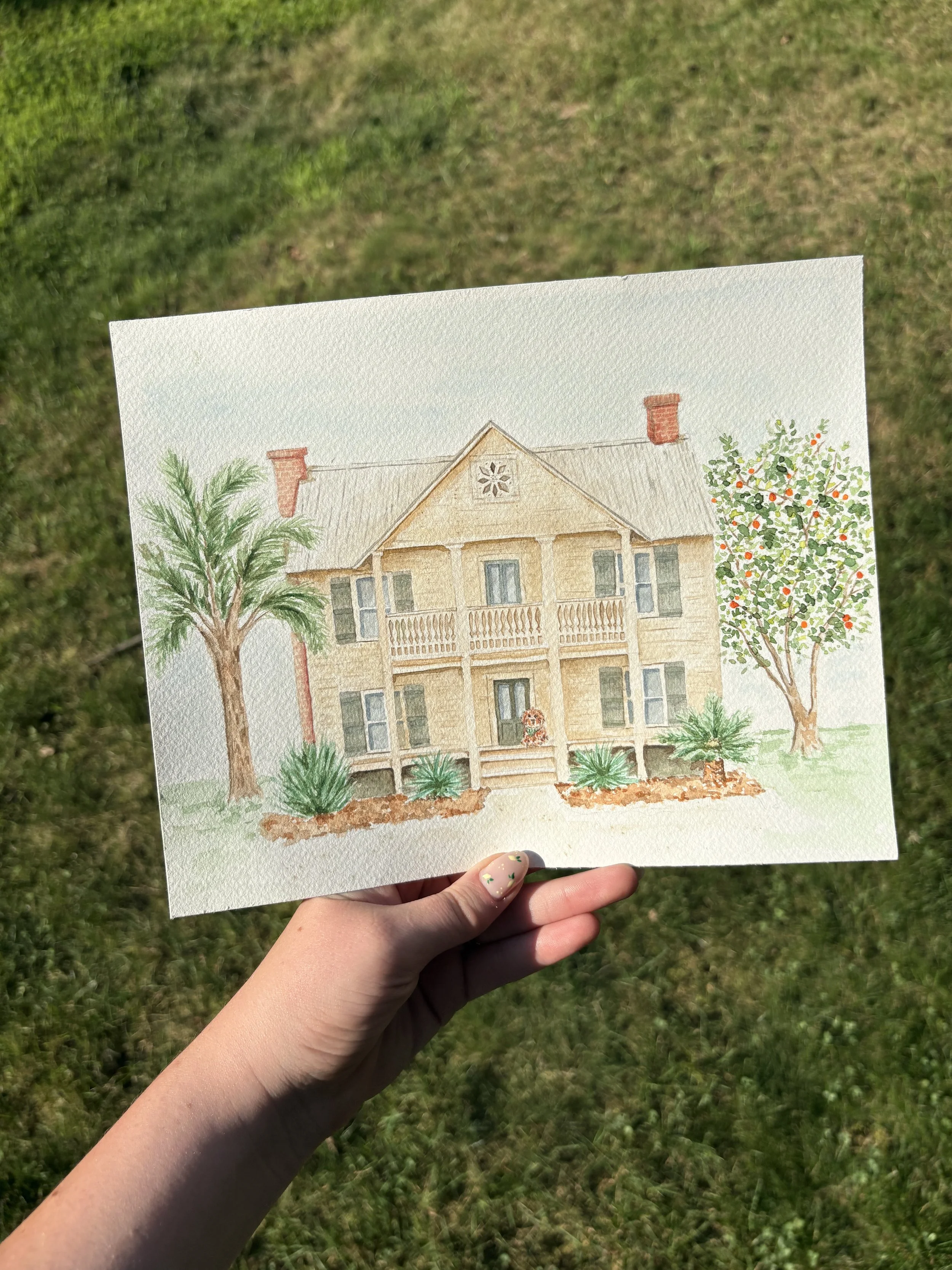 A hand holding a watercolor painting of a house with trees and plants, with a grassy lawn in the background.