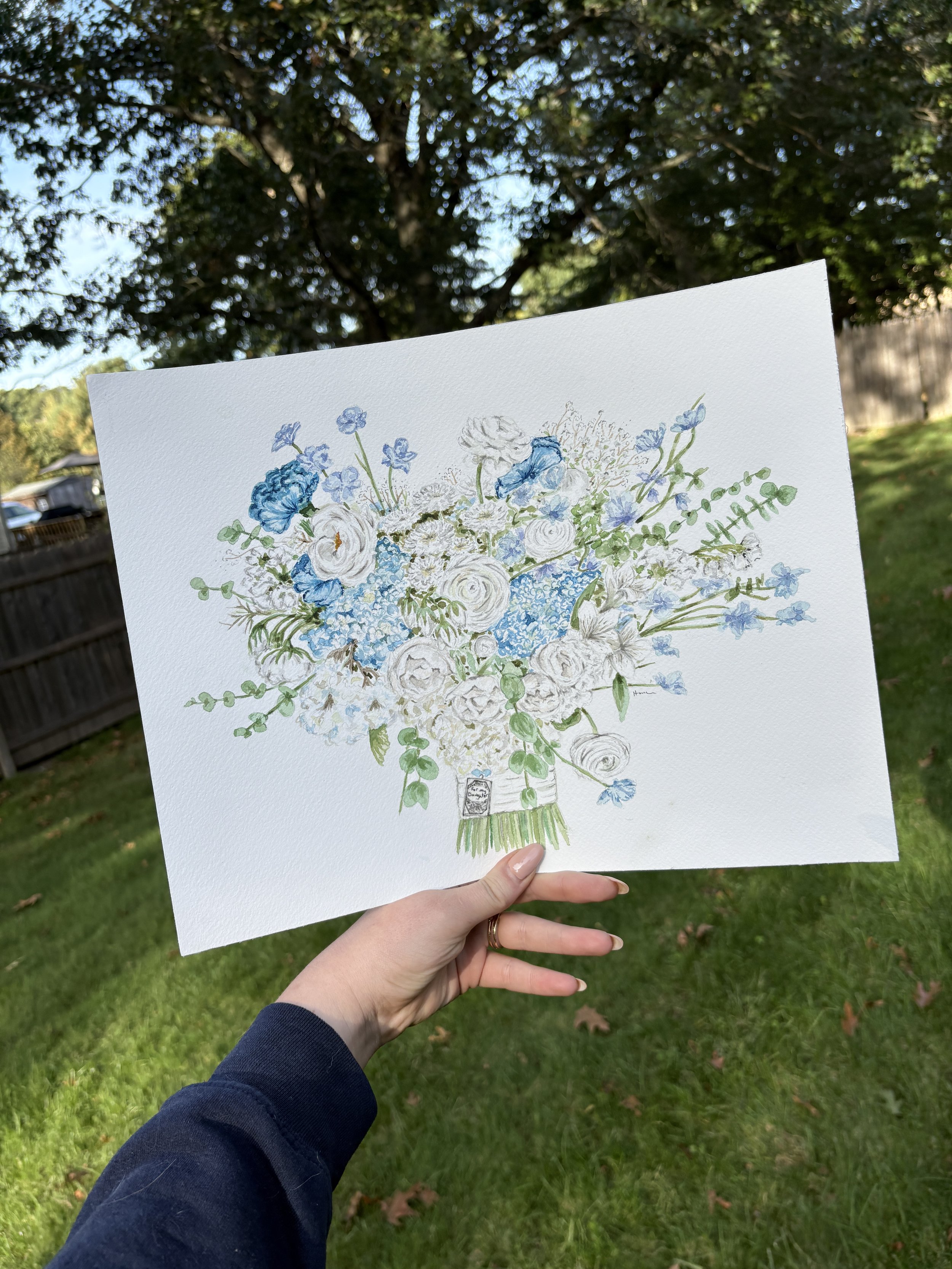 A person holding a watercolor painting of a bouquet of white and blue flowers outdoors, with trees and a fence in the background.