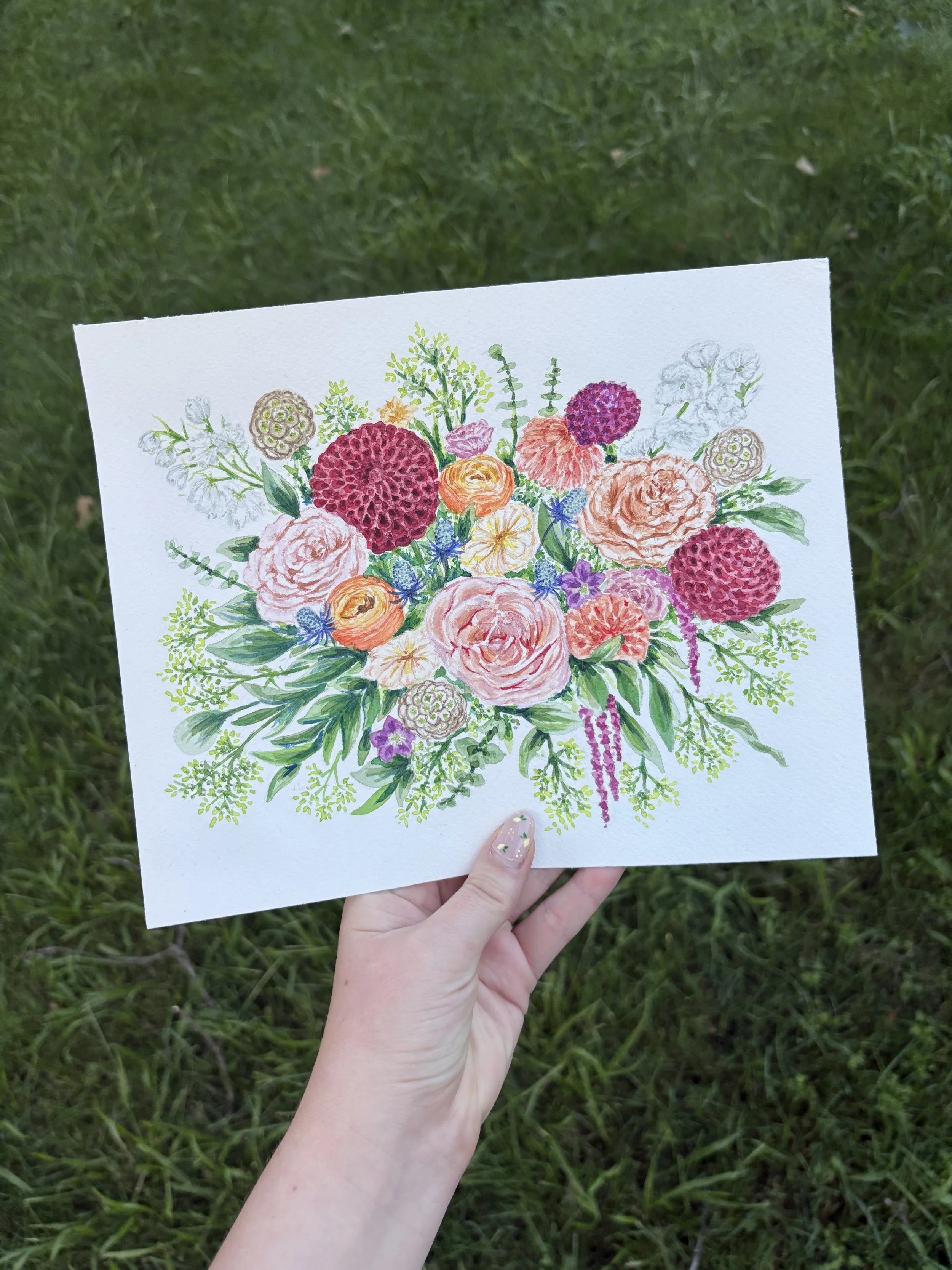 Hand holding a colorful floral watercolor painting against a grassy background.