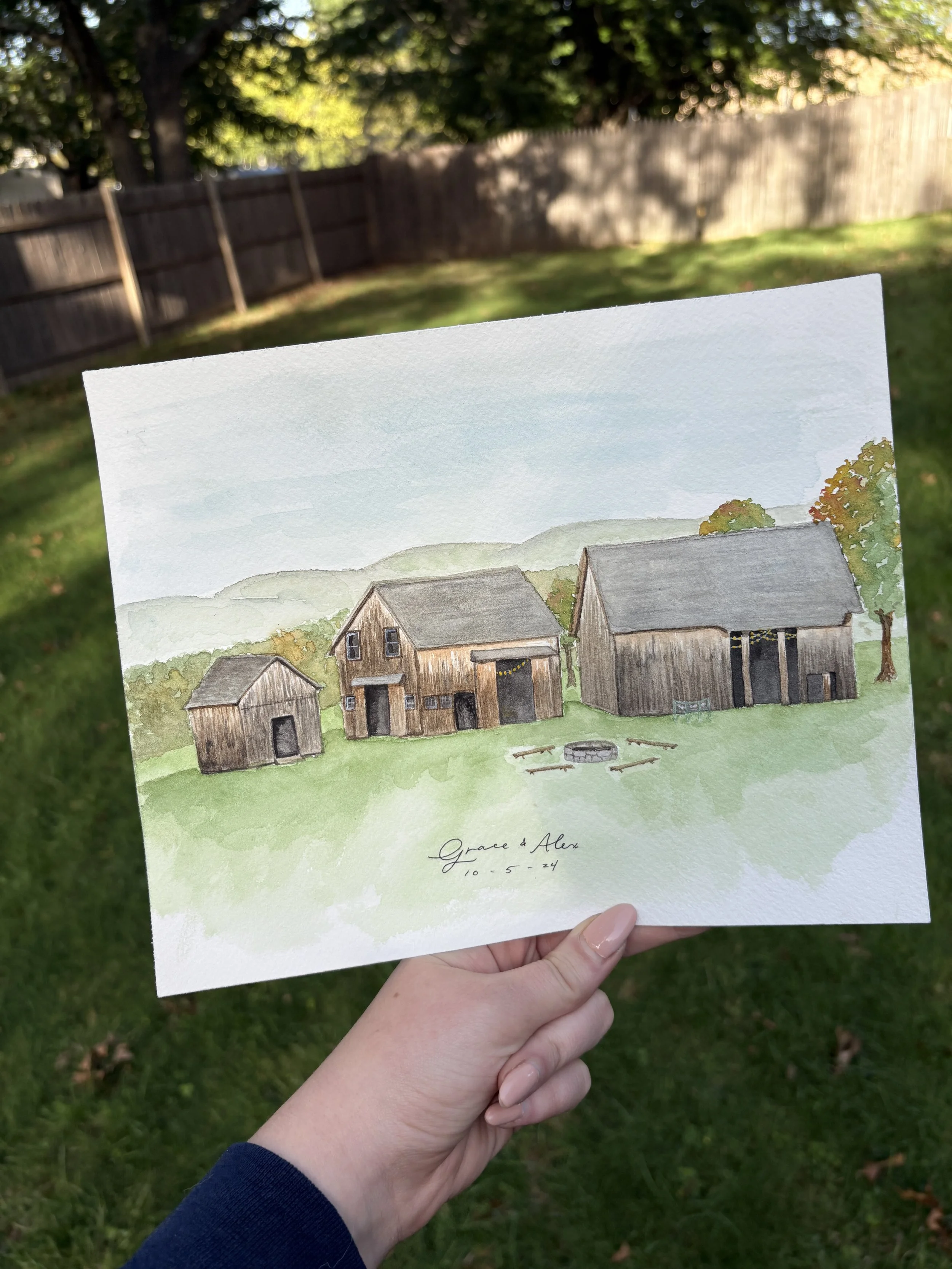 Watercolor painting of three wooden barns and a fire pit in a grassy yard, with trees and hills in the background, held by a person outdoors.