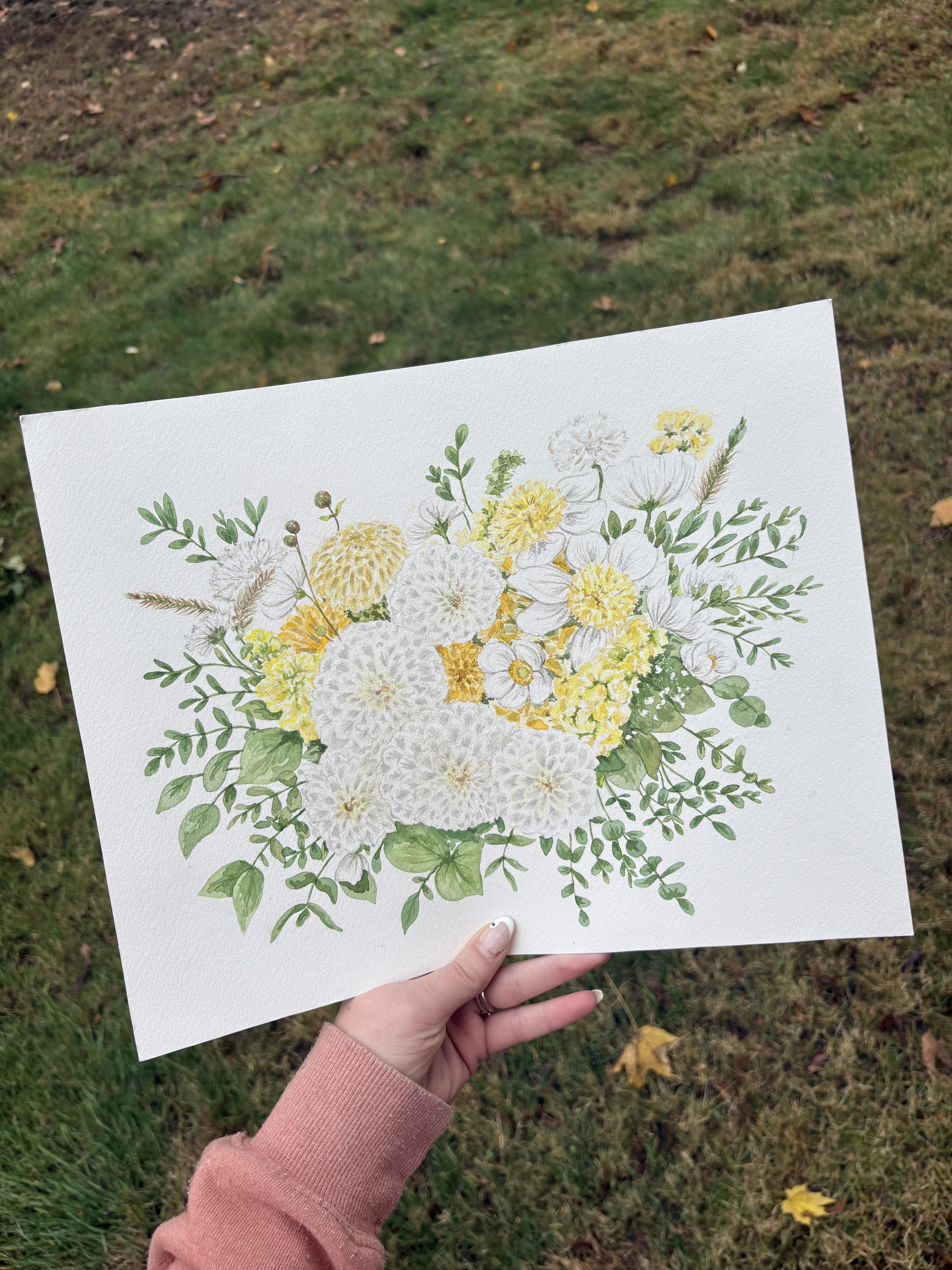A hand holding a white paper with a watercolor illustration of a floral bouquet featuring white, yellow, and green flowers and leaves, outdoors on grass.