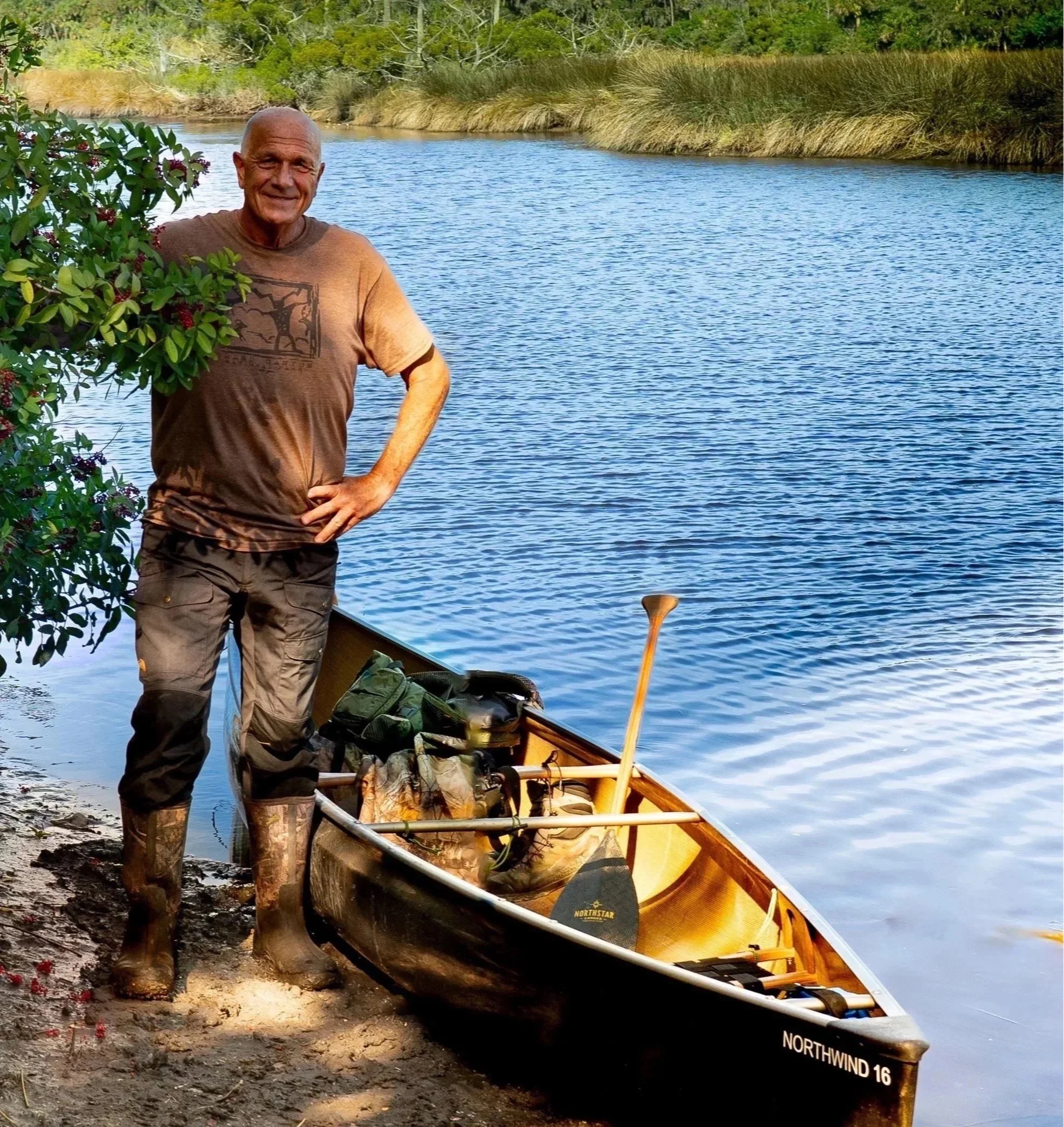 A man in outdoor gear stands next to a canoe on the riverbank, smiling at the camera, surrounded by nature.