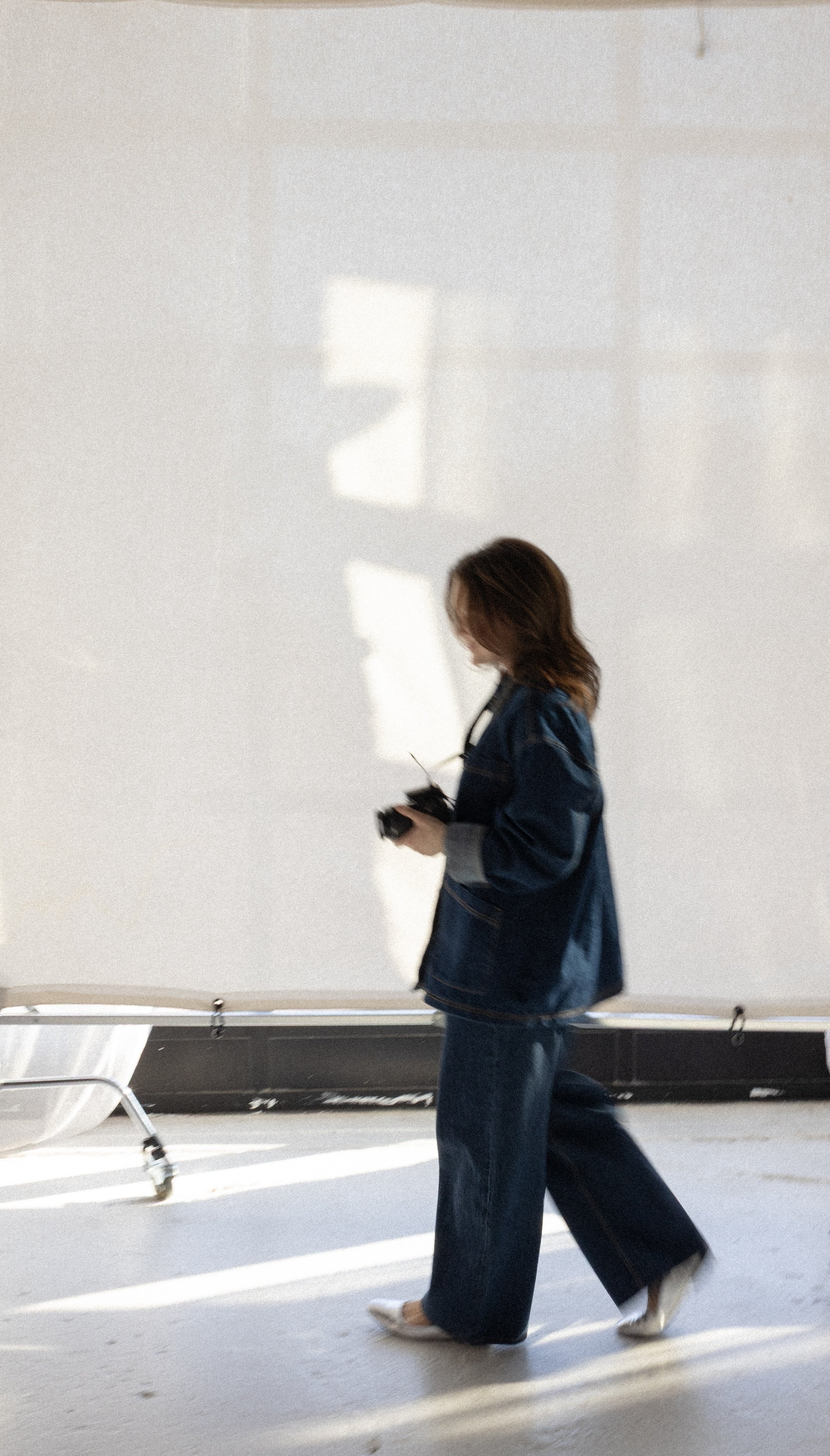Blurred person in denim outfit holding a camera, walking in front of a white backdrop in a brightly lit room.