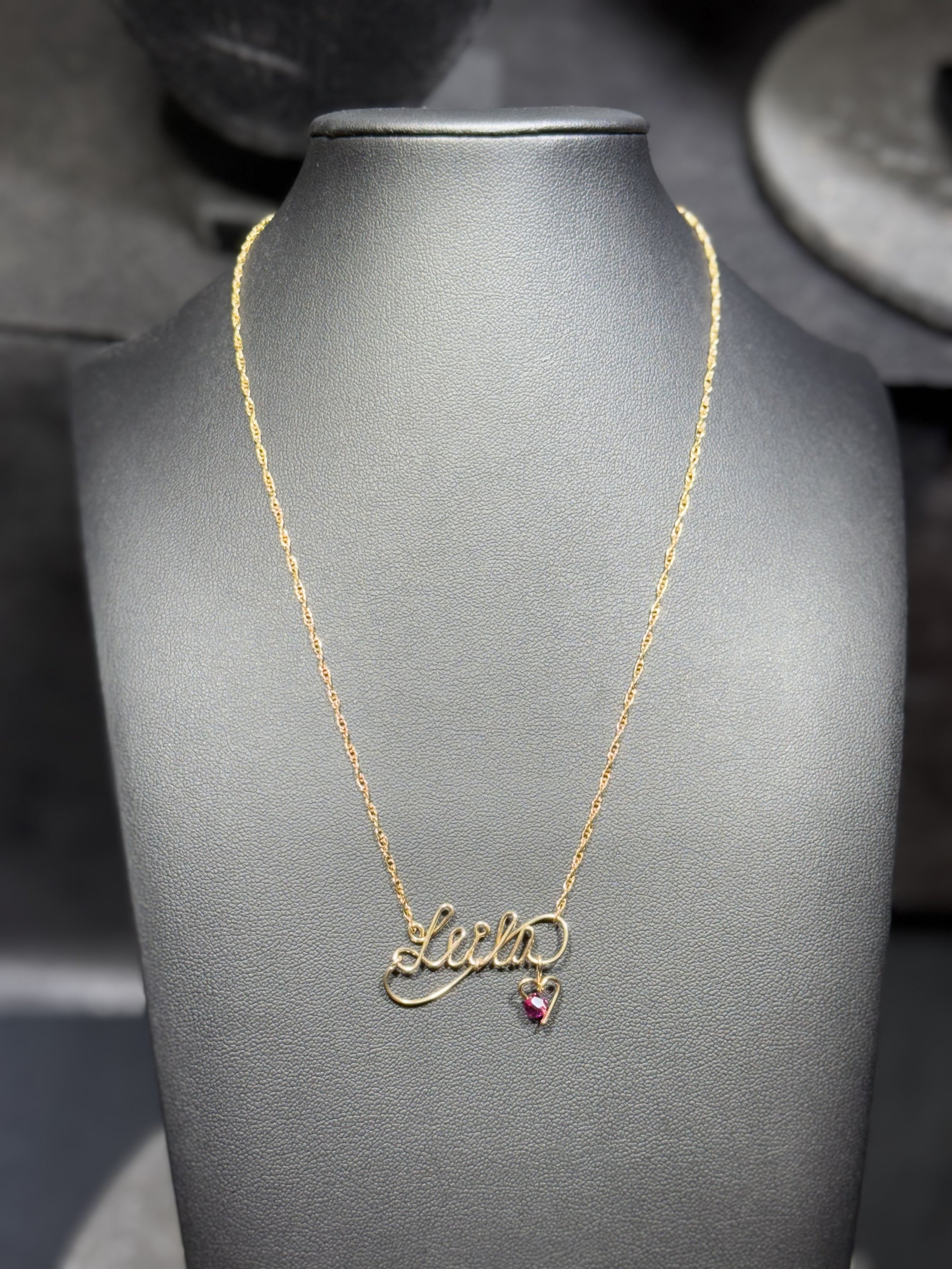 Gold necklace with a pendant spelling 'Lena' and a small pink heart charm, displayed on a gray jewelry stand.