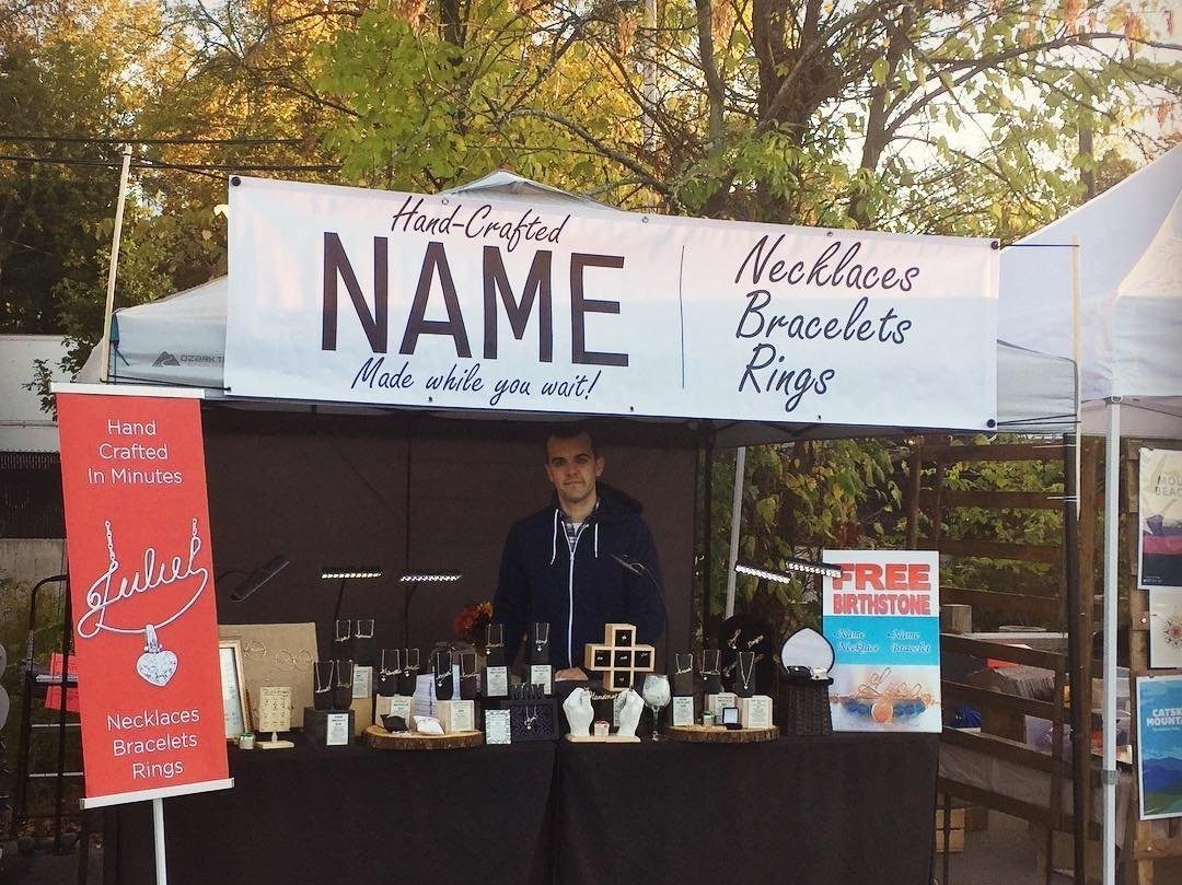 A booth selling handmade jewelry, including necklaces, bracelets, and rings, with a person standing behind the counter. The booth has a large sign advertising custom-made items and a smaller sign offering free birthstones.