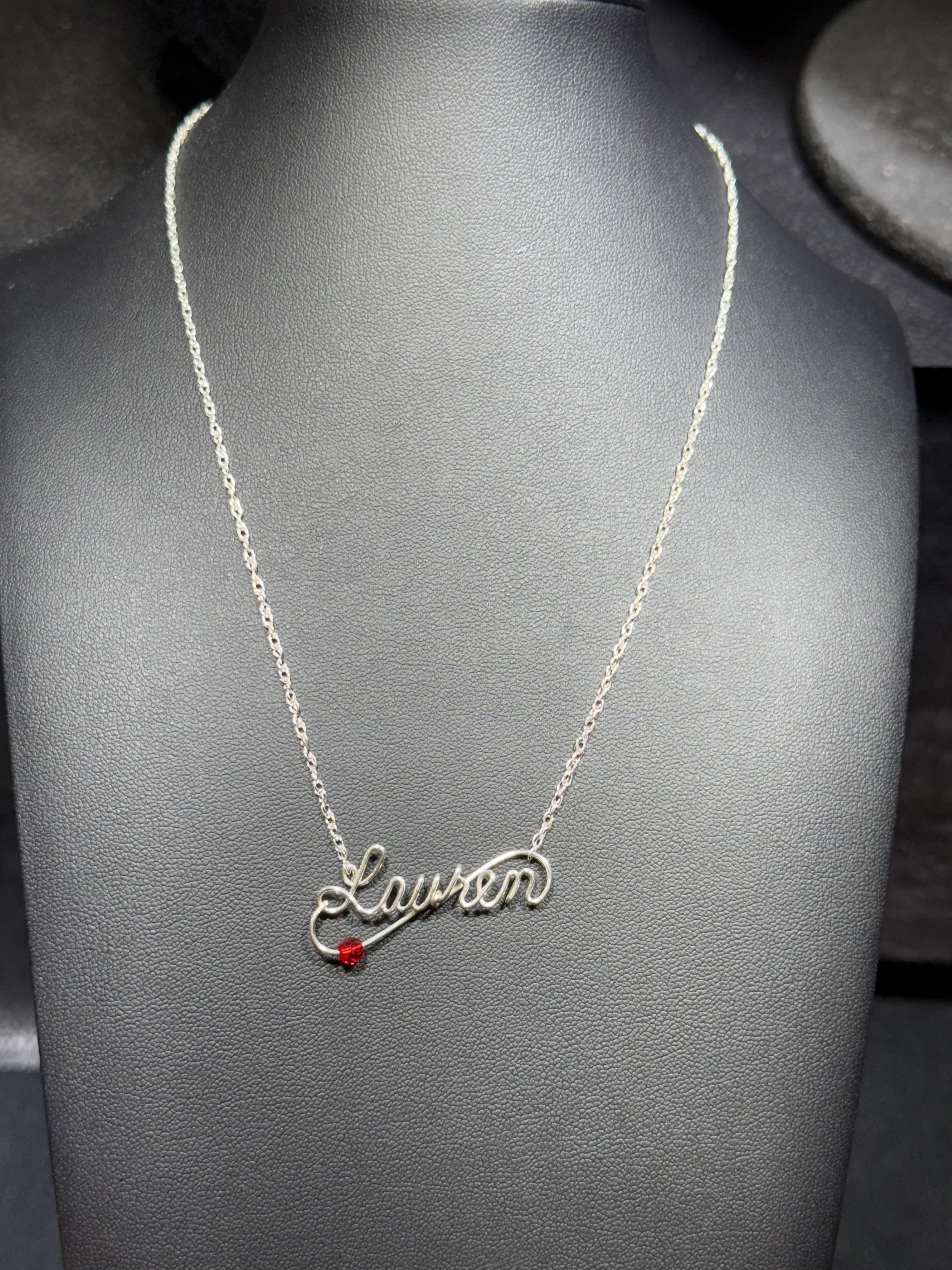 Silver necklace with a pendant that spell out the name "Lauren" and features a small red gemstone at the beginning of the name.